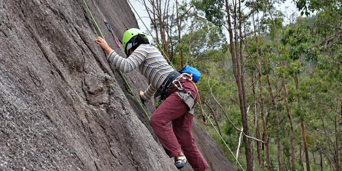 A climber in a green helmet moving up a steep granite slab on a top rope, with gum trees behind.