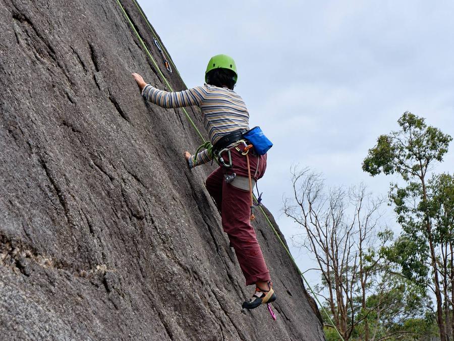 A climber in a green helmet and striped top moving up a granite slab, feet on the rock and the rope running up beside them.