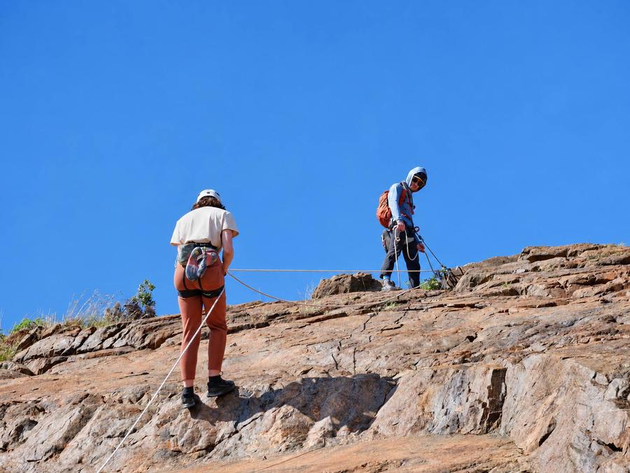 A climber and Evan near the top of a sunny rock face under a clear blue sky, a rope running between them.