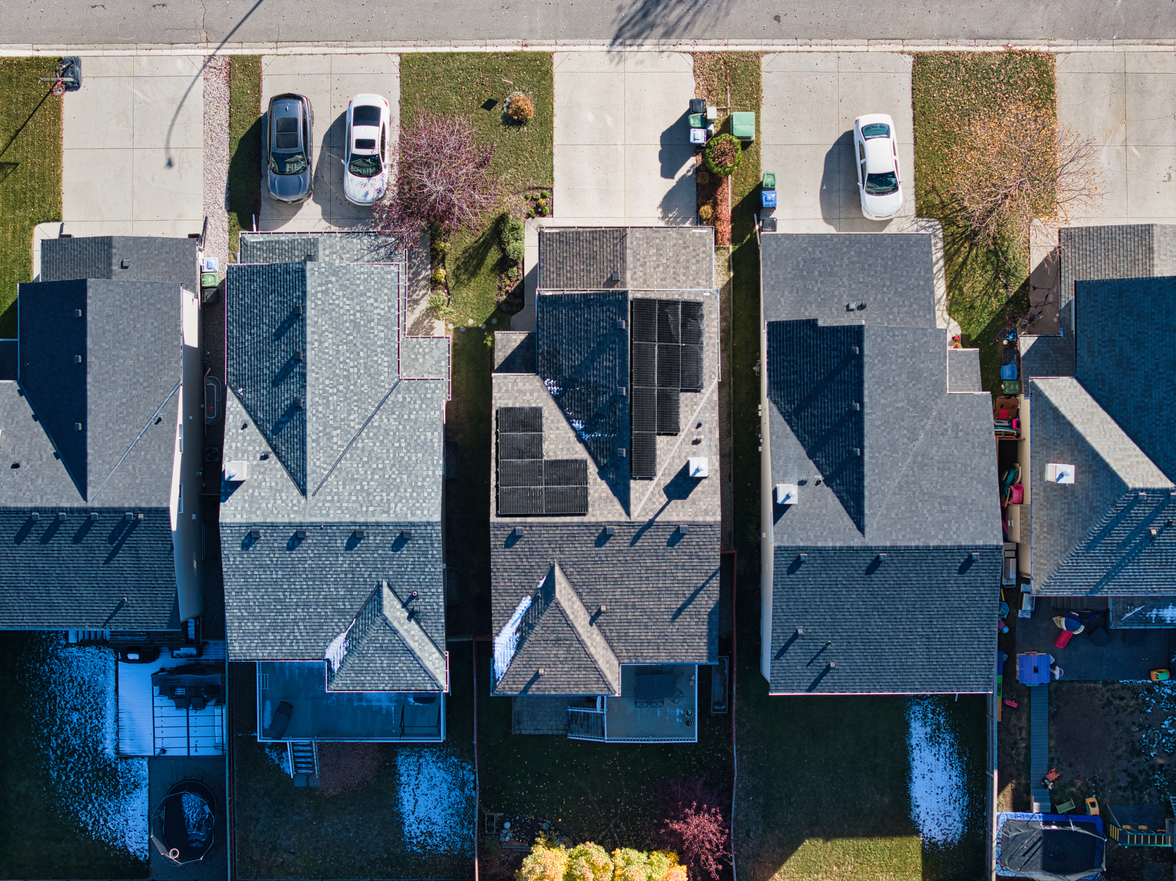 An aerial, top-down drone photograph of suburban houses, featuring one central home with modern black solar panels installed across two sections of its grey shingled roof.