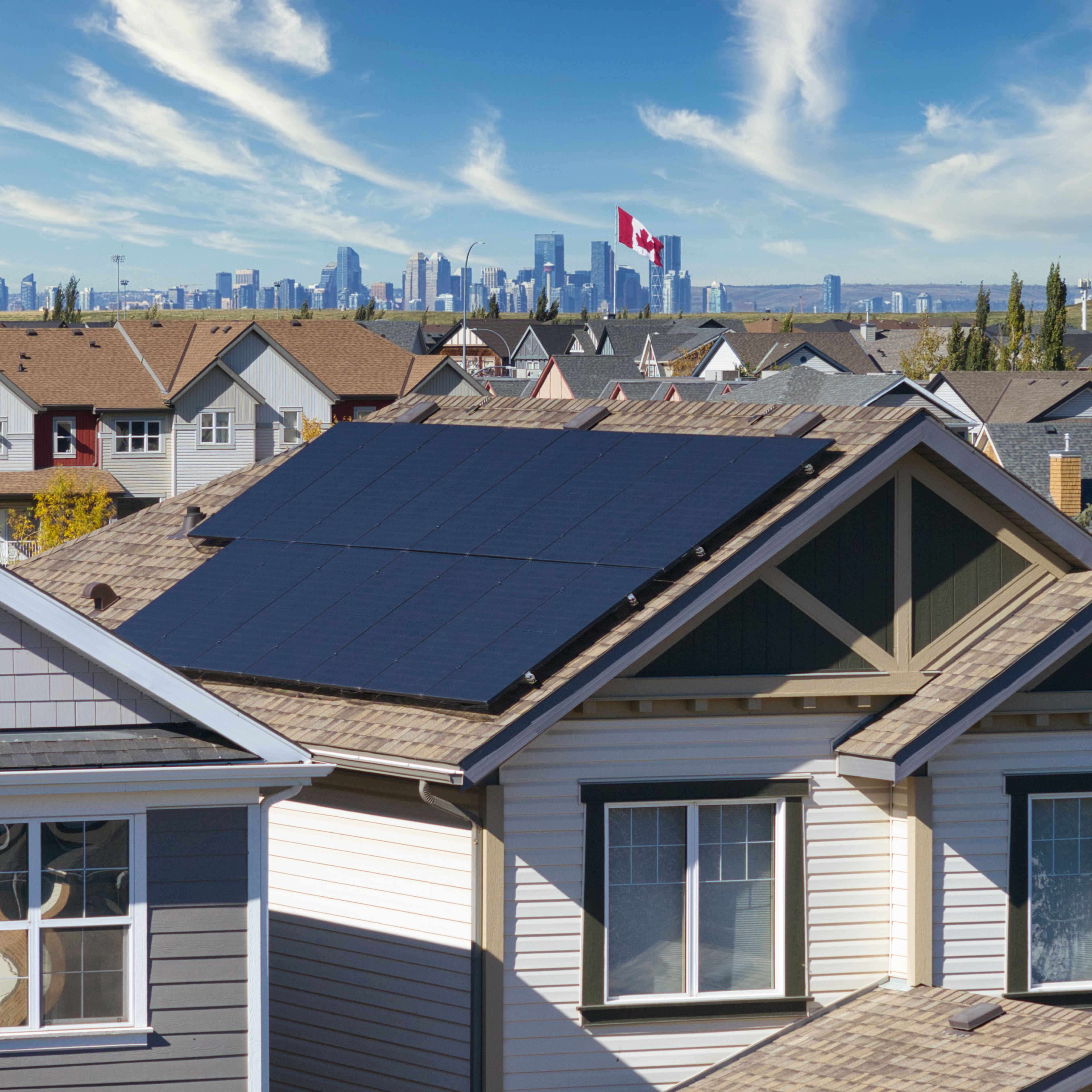 An elevated, close-up shot of suburban house rooftops, featuring a large array of dark solar panels installed on a sloped, shingled roof in the foreground. In the background, a sprawling city skyline stands under a bright blue sky filled with wispy white clouds, with a Canadian flag flying prominently on a tall flagpole in the distance.
