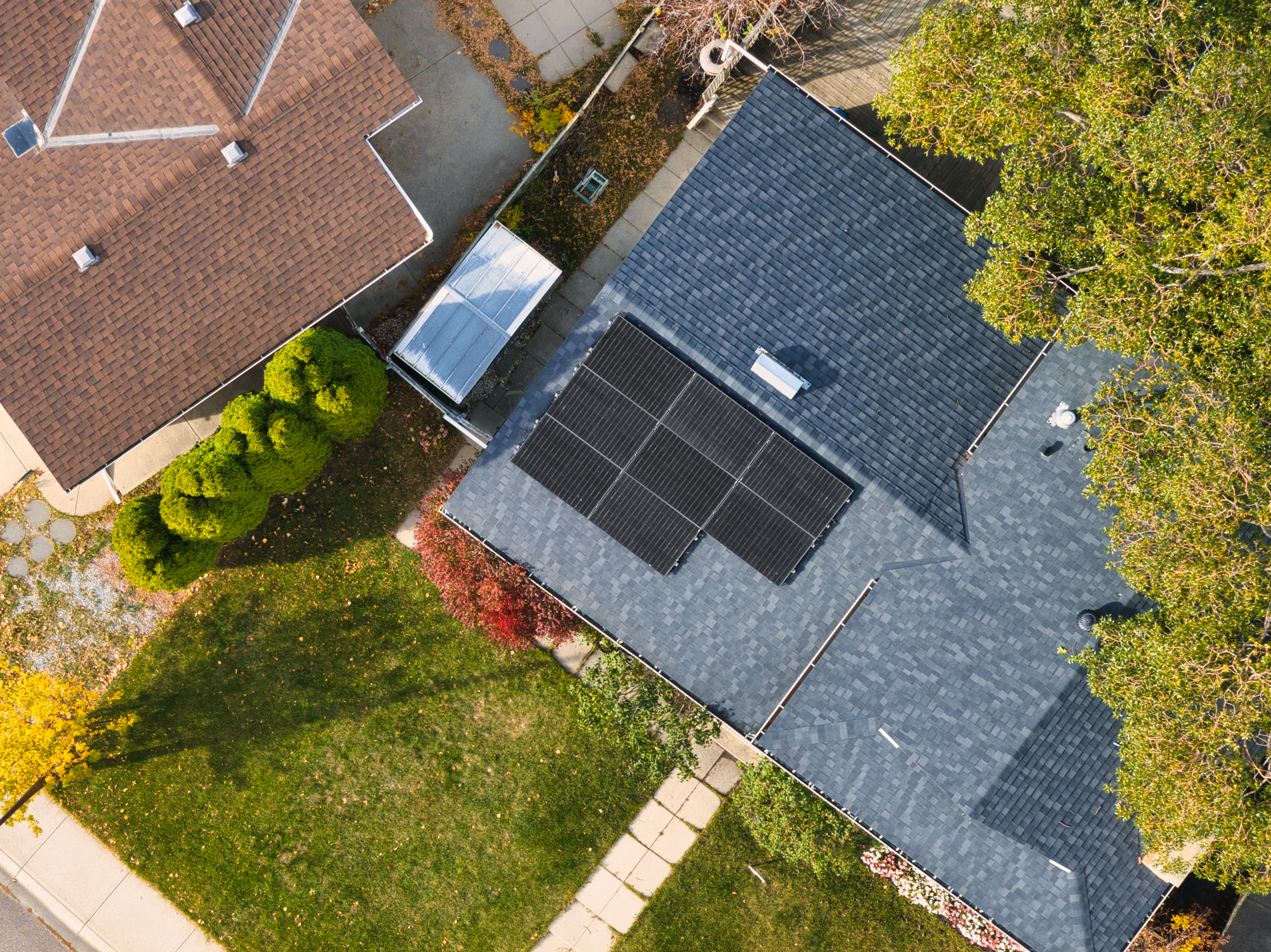 A top-down aerial drone photograph of a residential home featuring a neat array of six black solar panels installed on a dark grey shingled roof, surrounded by green lawns and autumn trees.