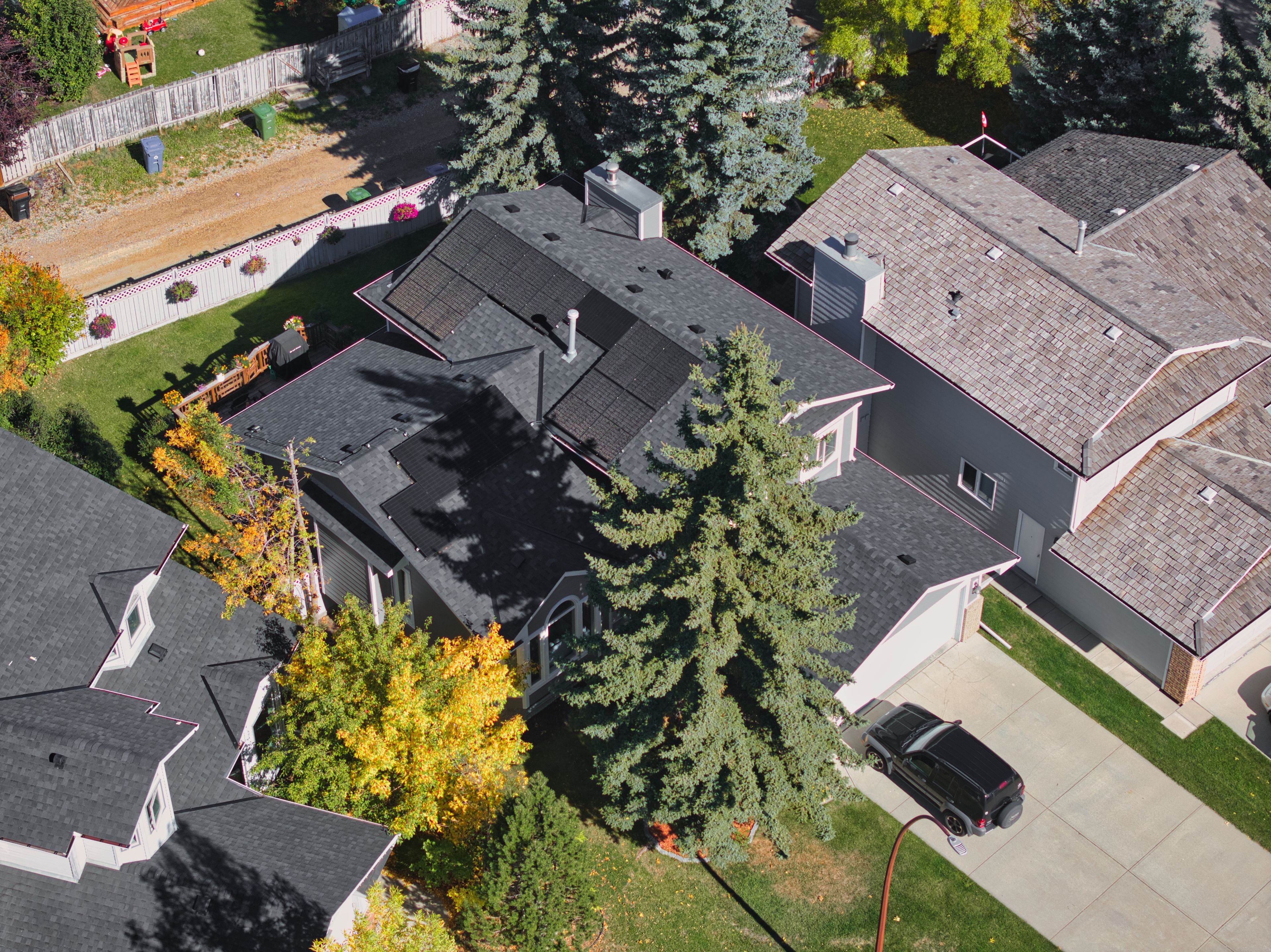 High-angle drone photograph of a home solar panel system installed by Firefly Solar on a multi-faceted dark asphalt shingle roof, strategically split to avoid shading from a large front-yard evergreen tree.