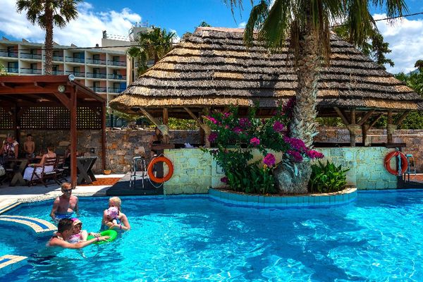 Oaspeți se relaxează la o piscină, cu un bar acoperit și palmieri, într-un peisaj însorit.