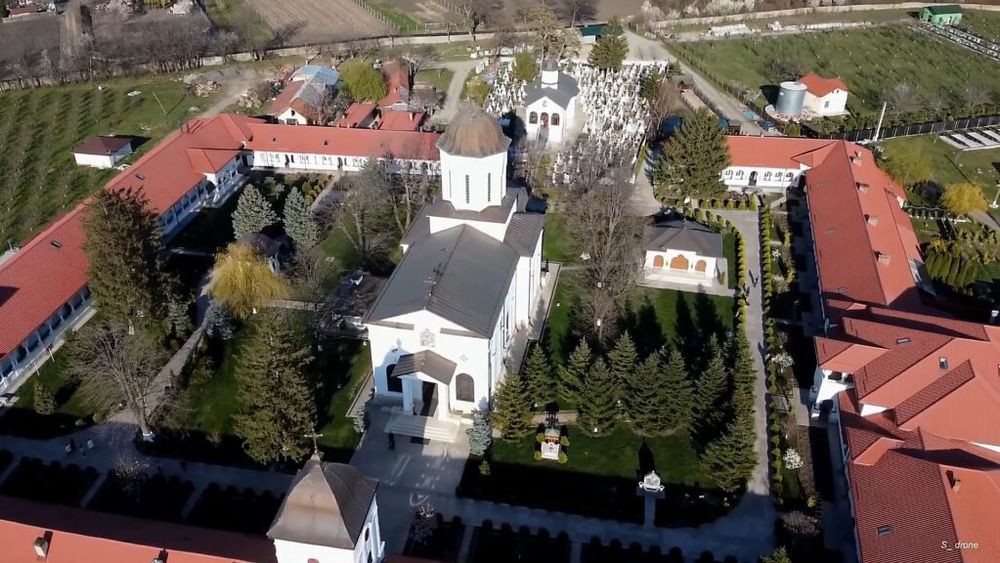 Mănăstire cu clădiri albe și acoperișuri roșii, înconjurată de arbori și un cimitir, pe fundal rural.