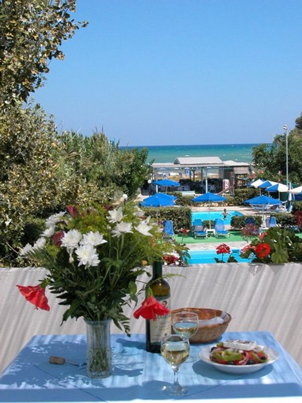 Masa cu flori, vin și mâncare, având în fundal marea și o piscină, sub un cer senin.