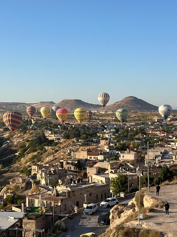 Balonase cu aer cald plutesc deasupra unui peisaj urban, cu case tradiționale și coline în fundal.