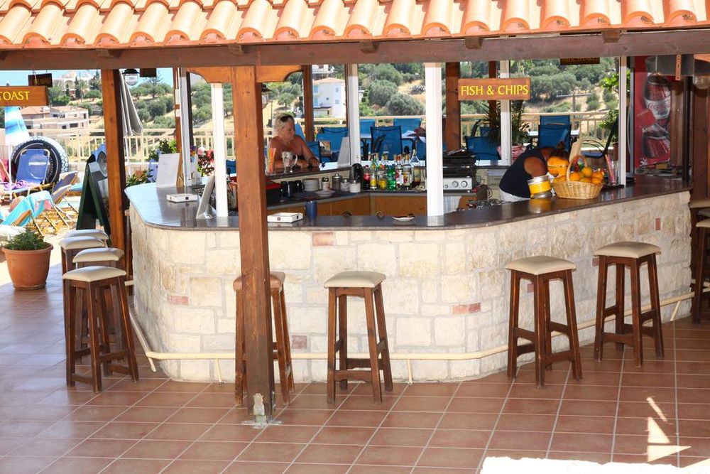 Bar exterior cu mesele de lemn, băuturi colorate și un meniu „Fish & Chips”, în fundal se văd piscine și peisaj.