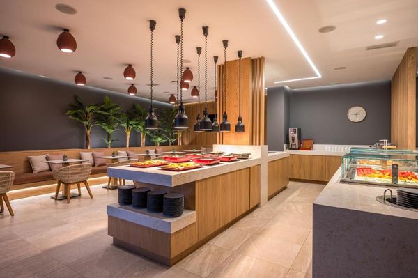 Spațiu modern de restaurant cu mese, plante, lumini suspendate și un bufet cu preparate diverse.