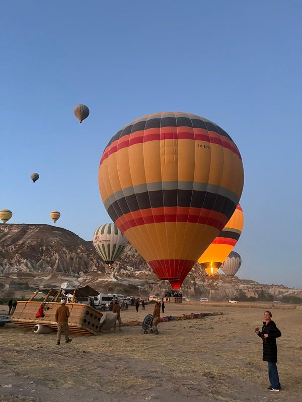 Colorful hot air balloons inflate in a clear sky, with people preparing for a flight in a scenic landscape.