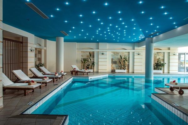 Piscină interioară cu apă cristalină, șezlonguri albe, plante decorative și tavan albastru cu lumini strălucitoare.