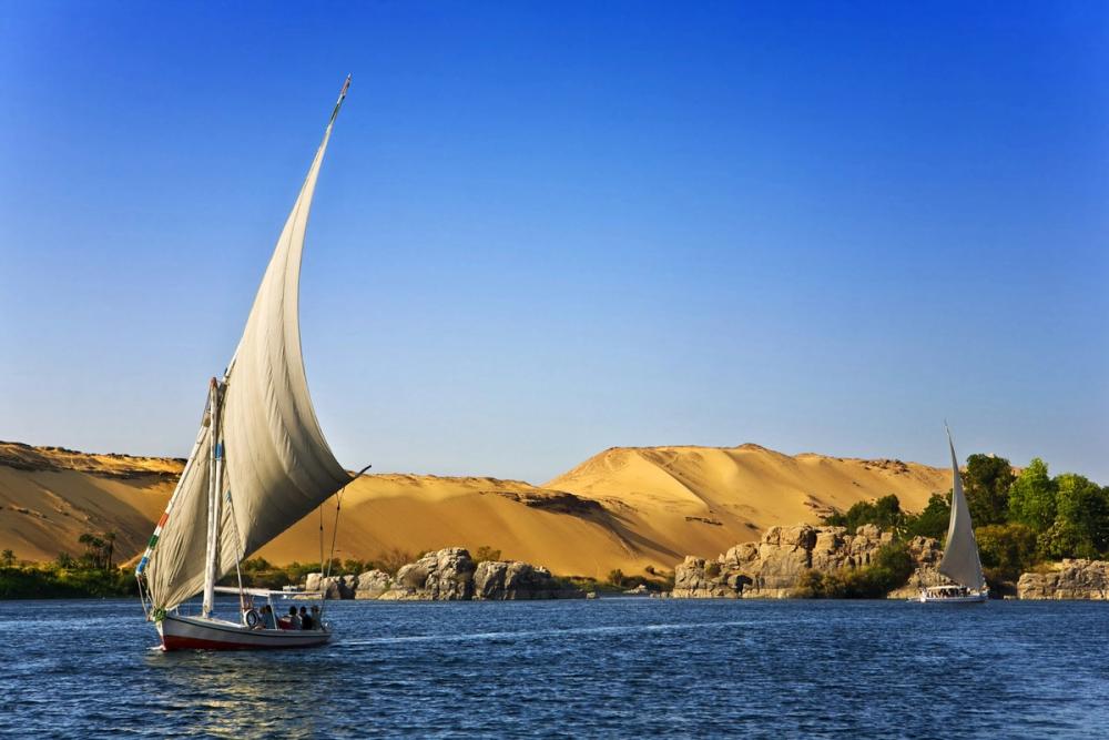Ambarcațiune tradițională felucca navigând pe Nil în Egipt, cu dune de nisip în fundal.