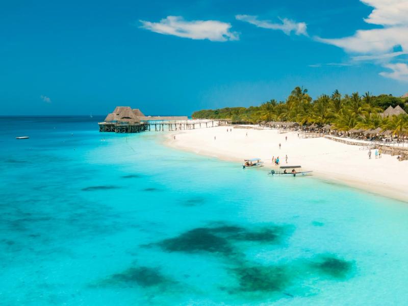 Plajă cu nisip alb și apă turcoaz în Zanzibar, Tanzania, cu bărci și palmieri tropicali.