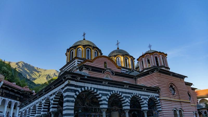Mănăstirea Rila din Bulgaria se înalță impunătoare la poalele munților, cu fațadele sale în dungi alb-negru și domurile elegante care strălucesc în lumina zilei. Arhitectura unică și peisajul montan din jur creează o atmosferă de liniște și spiritualitate profundă, specifică uneia dintre cele mai renumite mănăstiri ortodoxe din țară.