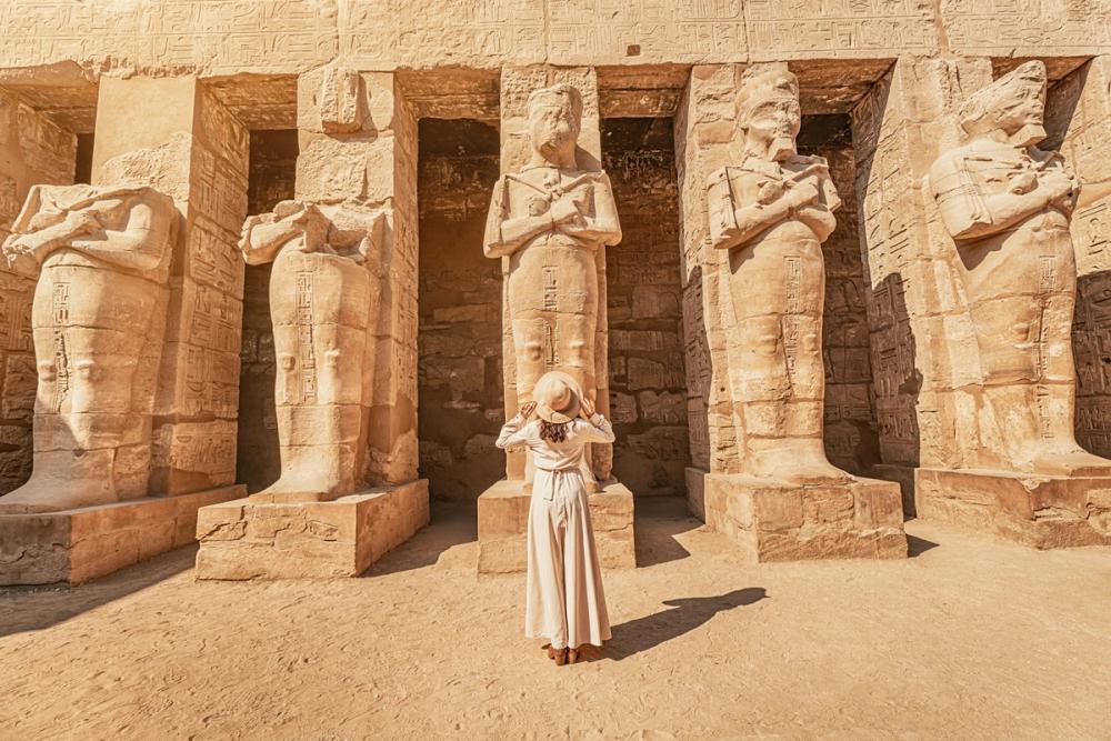 Turistă privind statuile antice dintr-un templu egiptean, înconjurată de hieroglife și piatră.
