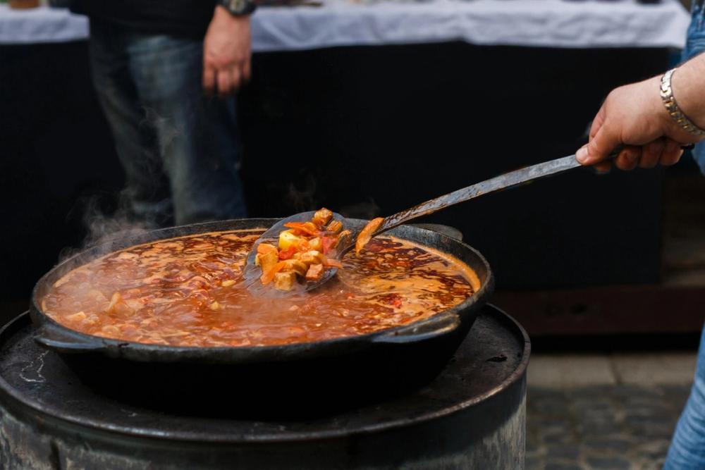 Mâncare tradițională românească gătită la ceaun în aer liber, cu legume și carne într-un sos roșu aromat, amestecată cu o lingură mare de metal.