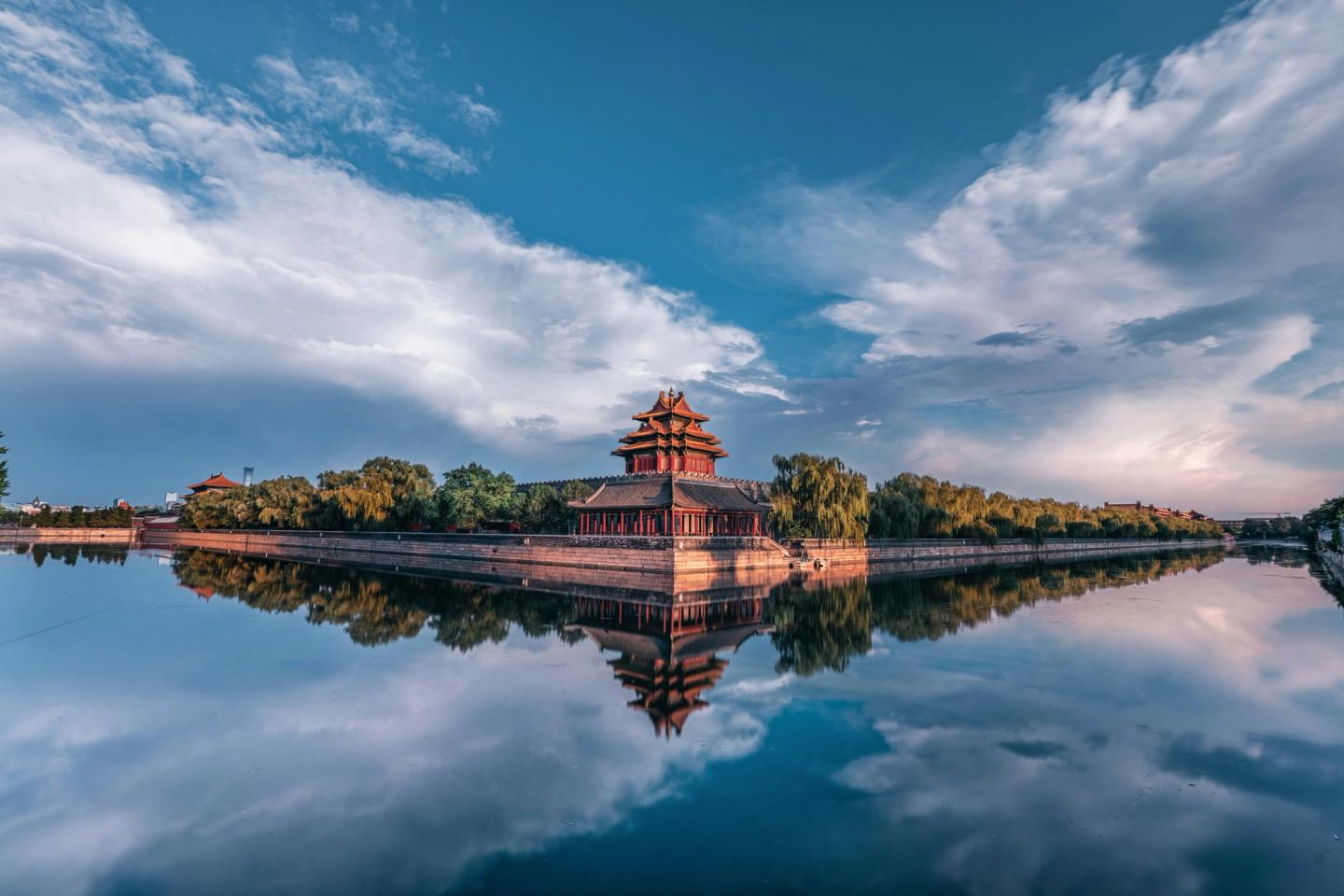 Pavilion chinezesc tradițional din Orașul Interzis reflectat în apele liniștite ale șanțului de apă, sub un cer parțial înnorat.