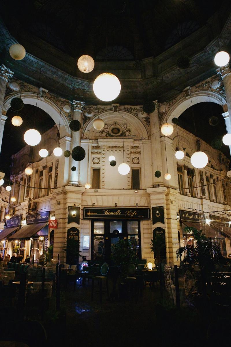 Cafenele și clădiri istorice luminate seara în Centrul Vechi din București, România, cu lumini suspendate și terase animate.