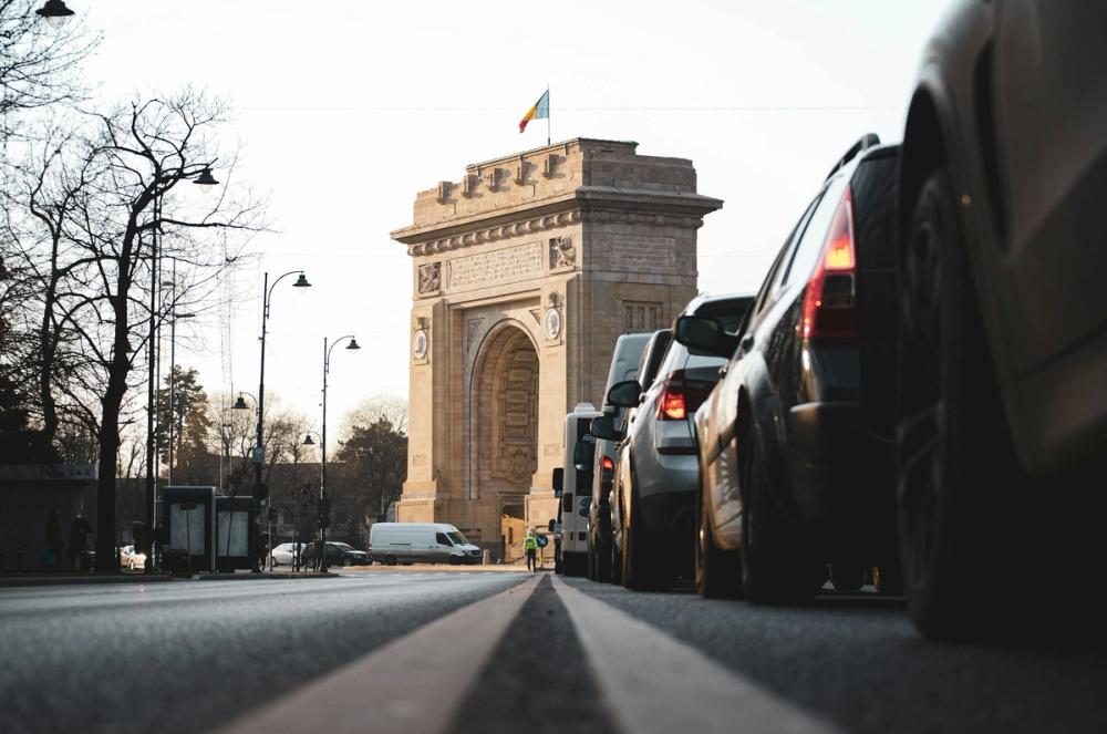 Arcul de Triumf din București, România, văzut de la nivelul străzii, cu mașini aliniate în trafic și steagul României arborat în vârf, într-o zi luminoasă de iarnă.