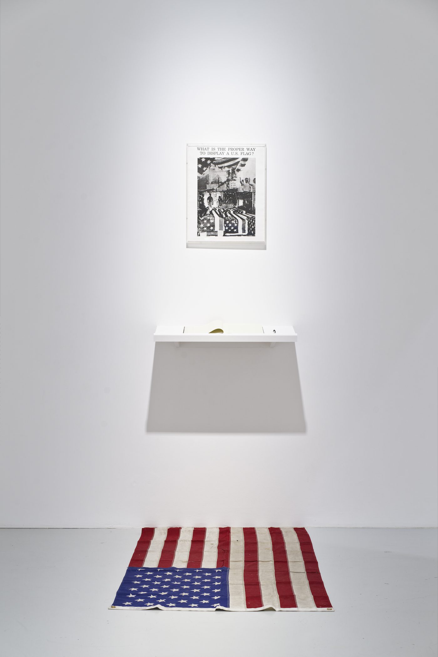 Dread Scott, What is the Proper Way to Display a US Flag?, 1988, installation with silver print, US flag, book(s), pen, shelf, active participation. Collection of the artist. © Dread Scott. View of the exhibition Globalisto. A Philosophy in Flux at the Musée d’art moderne et contemporain de Saint-Étienne Métropole, from June 25th to October 16th, 2022. Crédit photo : C. Cauvet / MAMC+