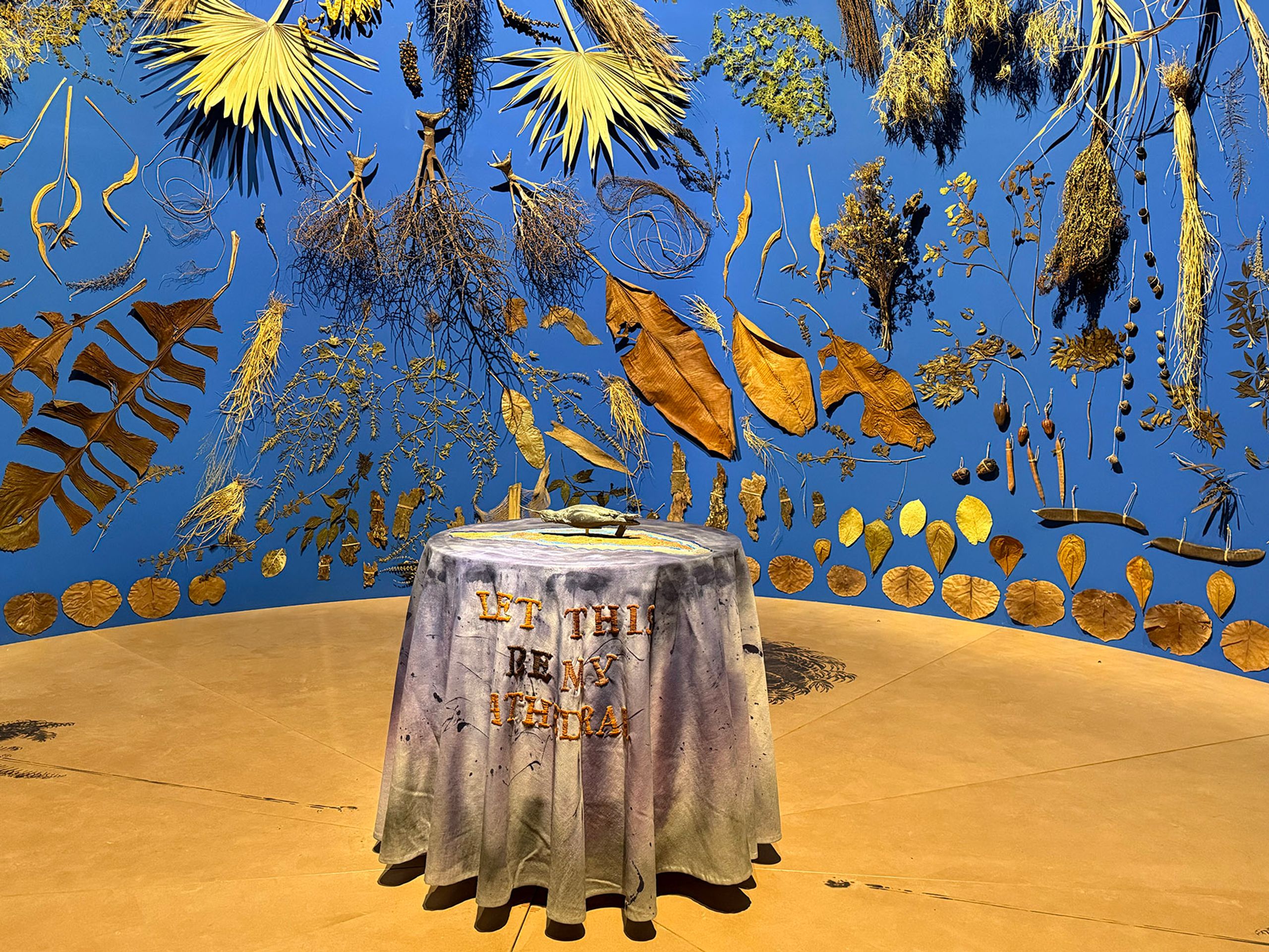 A blue wall decorated with various dried plants and leaves, with a table covered in a cloth reading "LET THIS BE MY ITHACA" and holding a small metallic boat in the foreground.