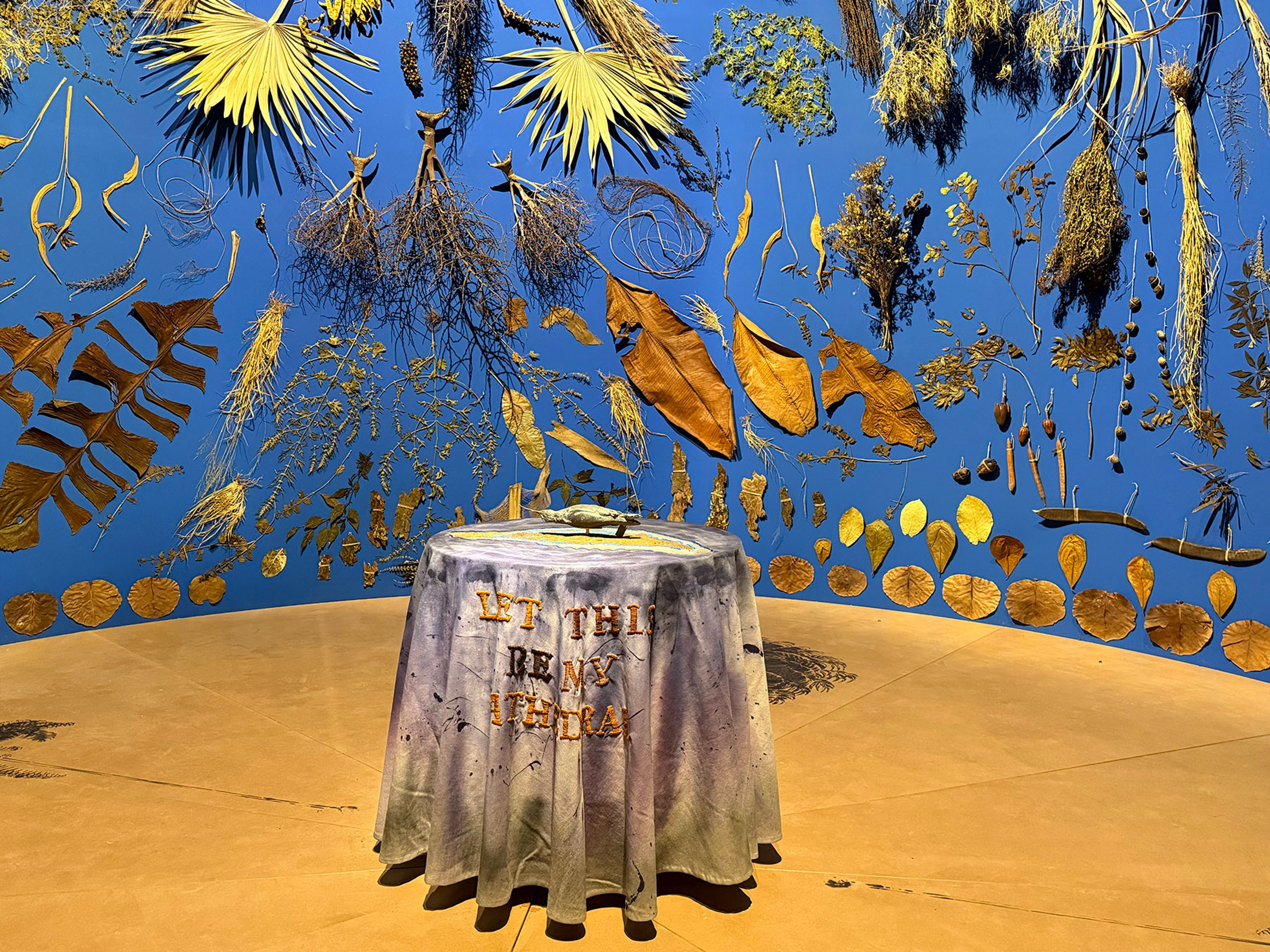 A blue wall decorated with various dried plants and leaves, with a table covered in a cloth reading "LET THIS BE MY ITHACA" and holding a small metallic boat in the foreground.