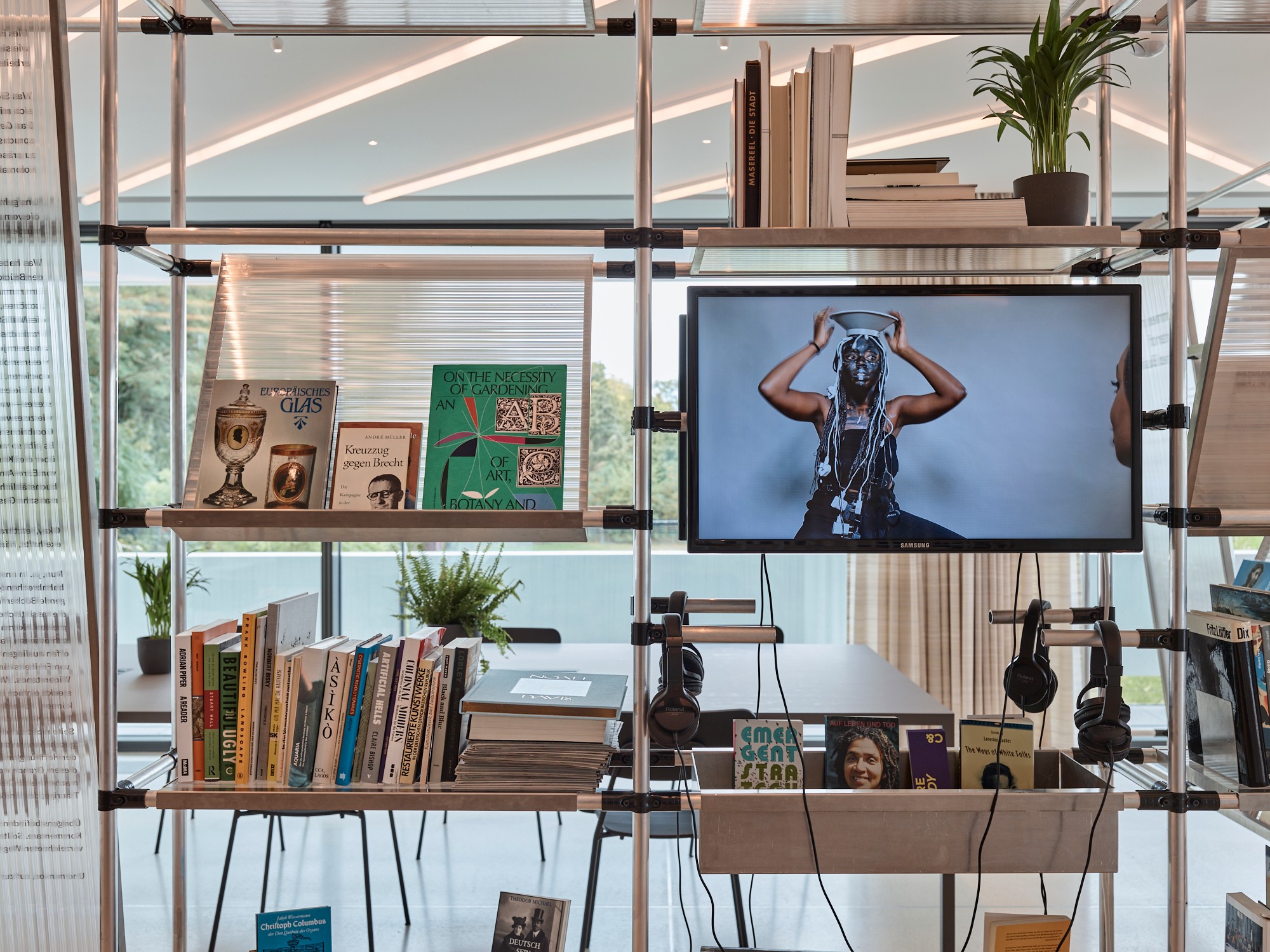 A modular shelving unit with books, plants, and headphones, features a TV showing a person balancing a bowl on their head, set against large windows.