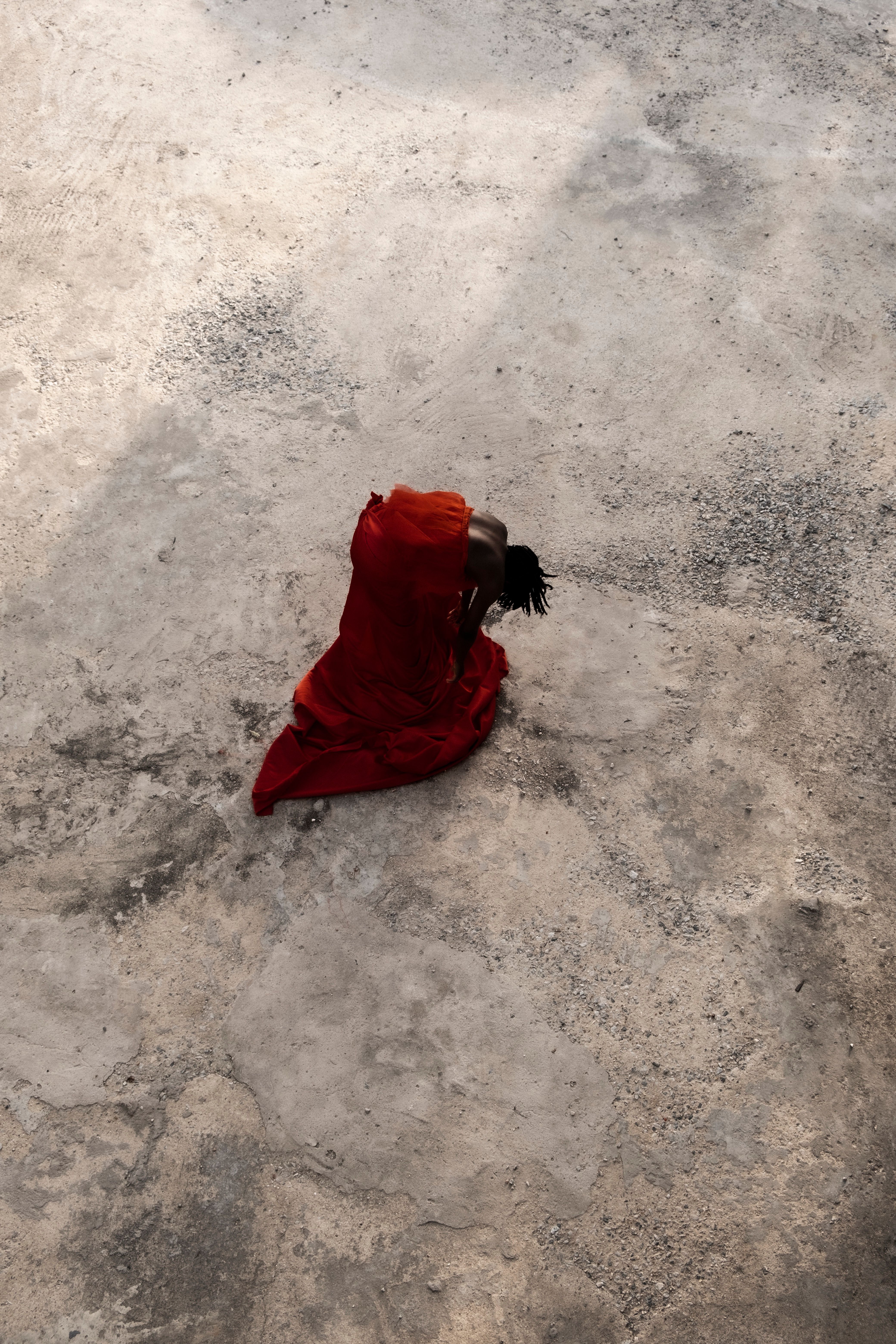 Overhead view of a figure in a flowing red garment bent over on a textured grey ground.