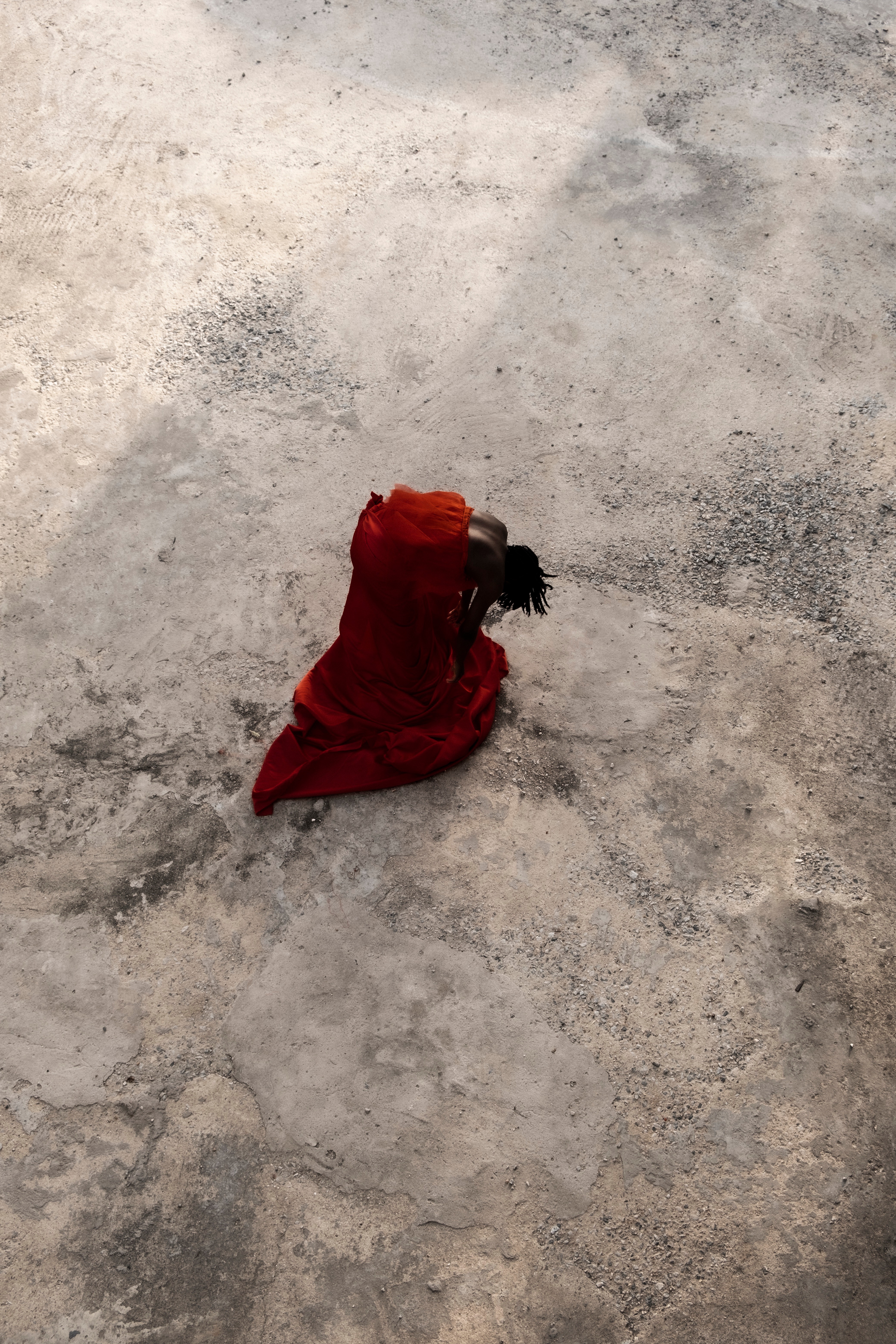 Overhead view of a figure in a flowing red garment bent over on a textured grey ground.