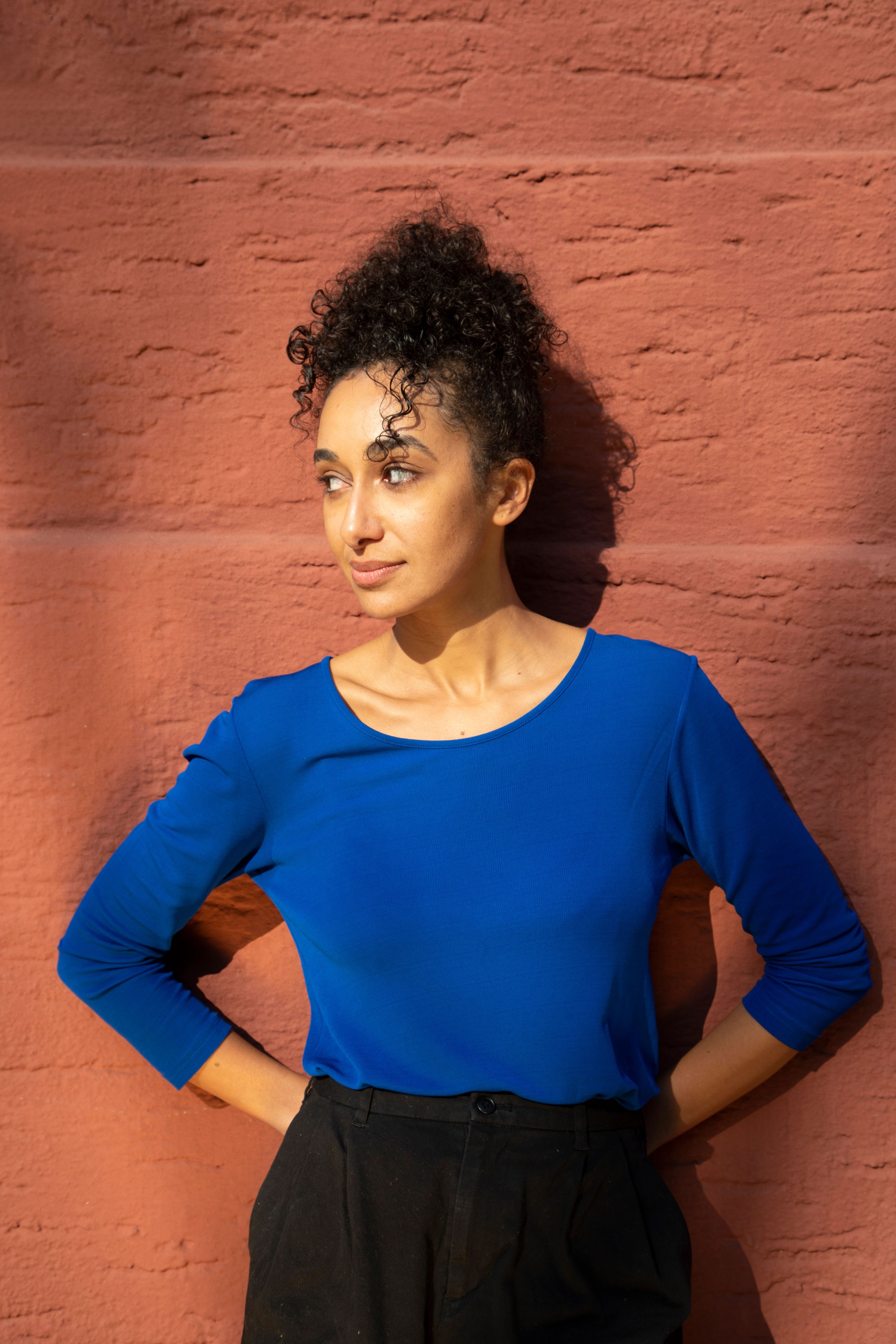 A woman with dark curly hair in a blue shirt and black pants stands against a red brick wall, looking left with hands on her hips.