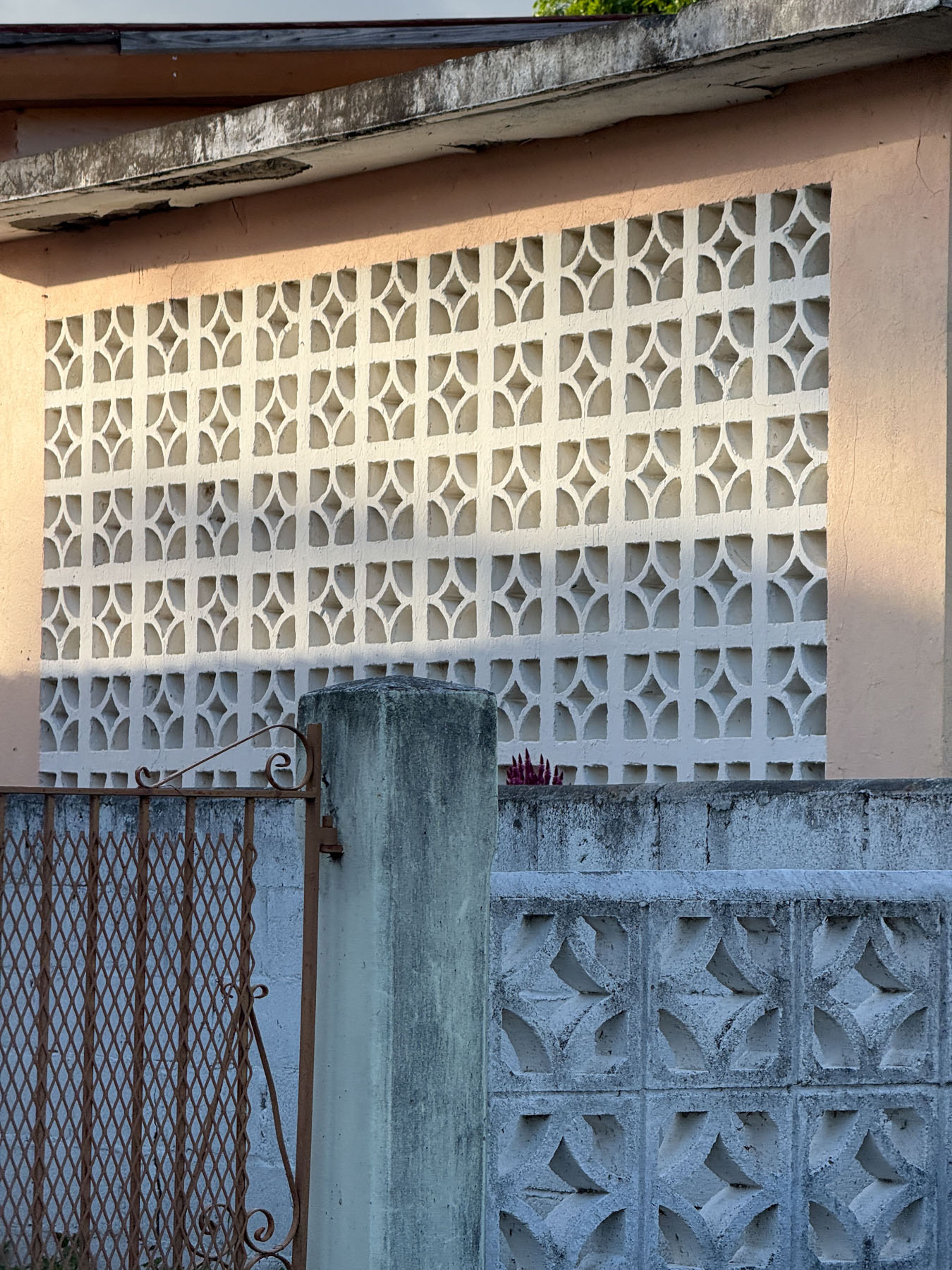 A pink building wall with a large section of white decorative concrete blocks, a rusty gate, and a lower grey patterned concrete block wall.