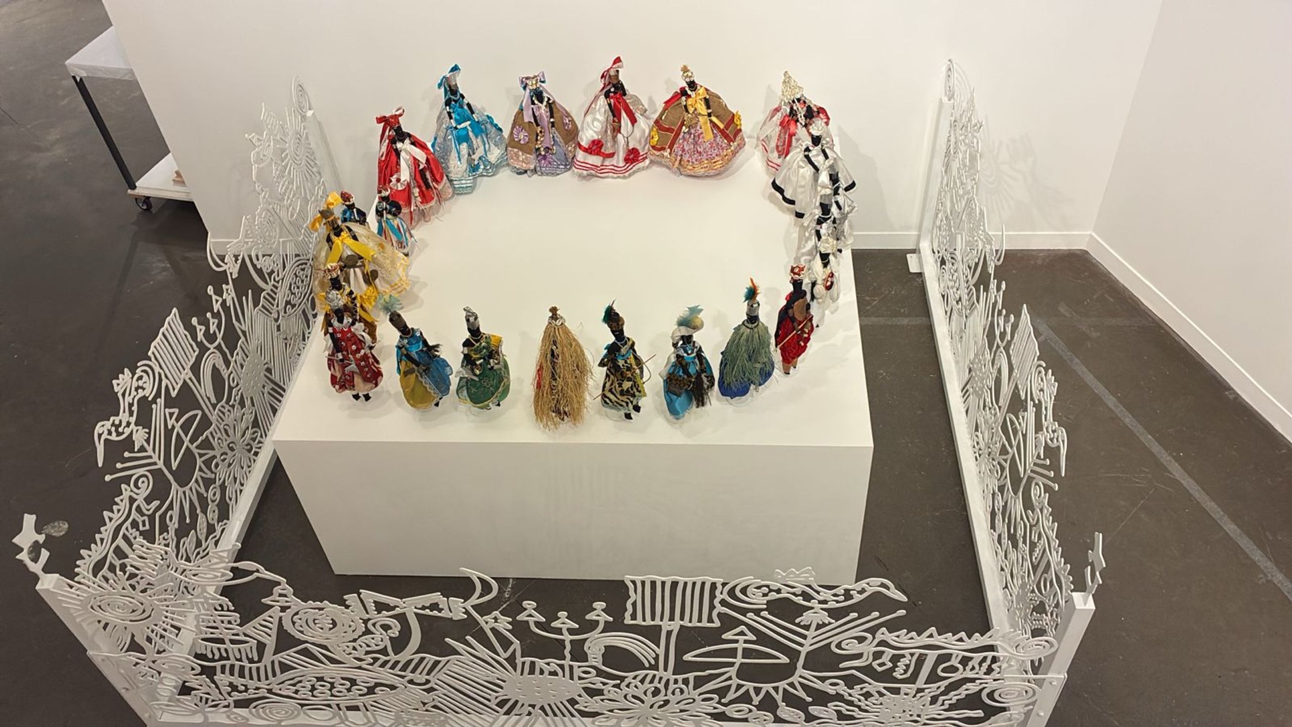 Colorful figures in elaborate dresses stand on a white pedestal, framed by intricate white screens in an art gallery.