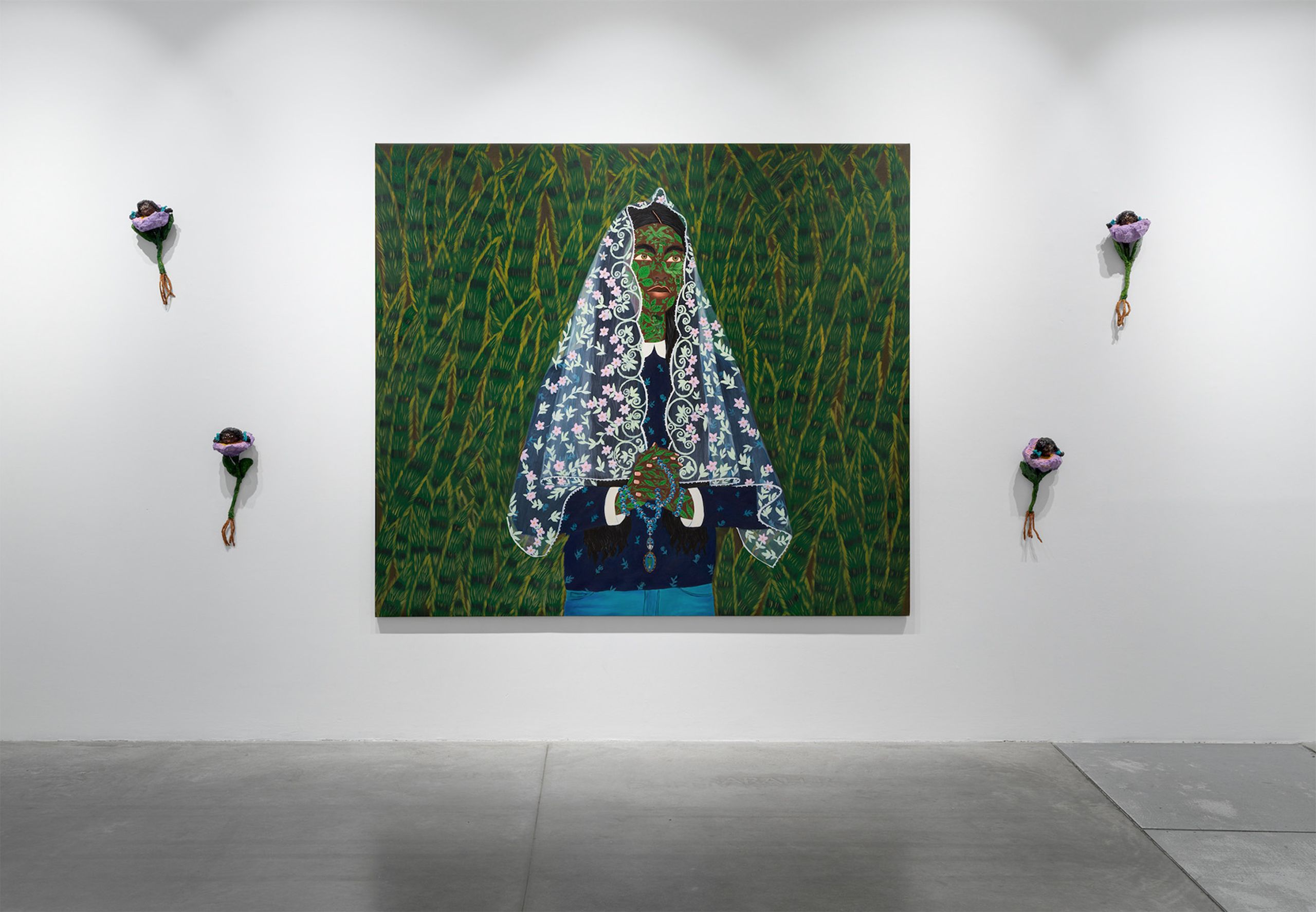 A large painting of a green-skinned figure in a patterned blue and white veil, holding a rosary, against a green background, is flanked by four purple flower sculptures with hanging roots on a white gallery wall.