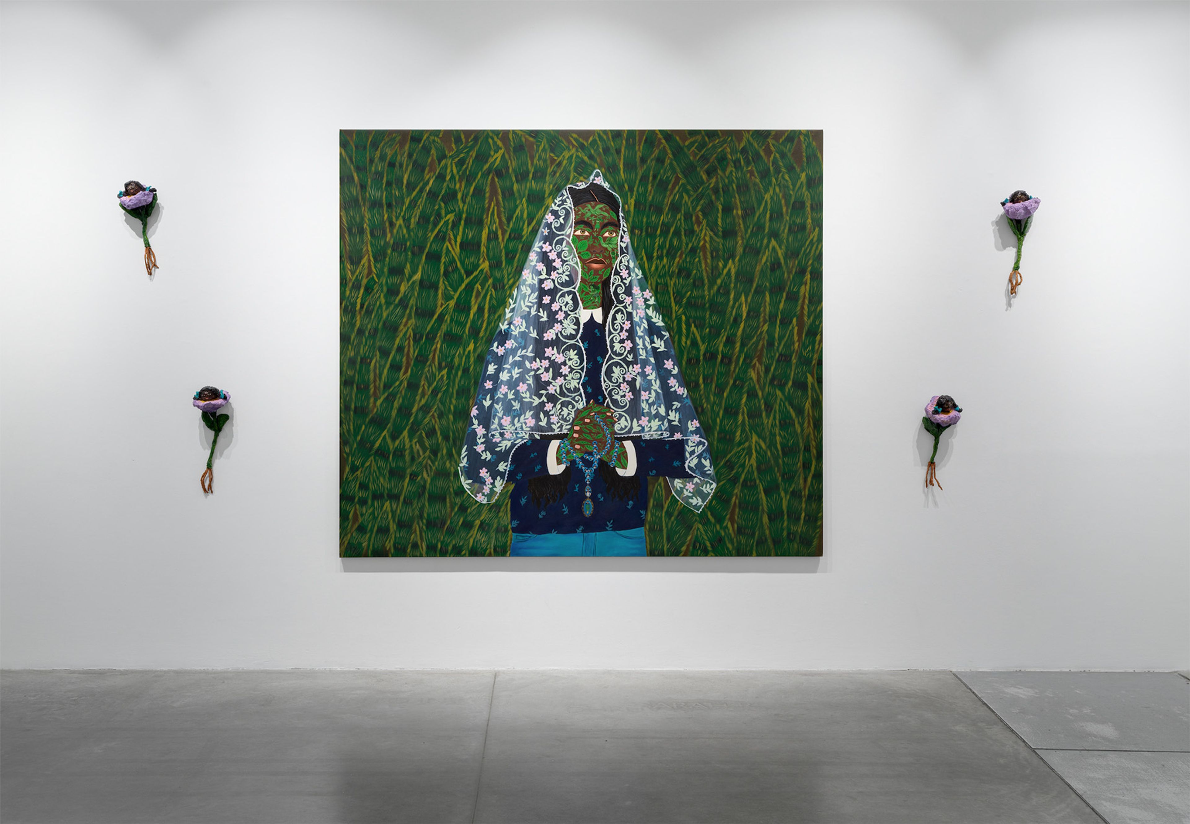 A large painting of a green-skinned figure in a patterned blue and white veil, holding a rosary, against a green background, is flanked by four purple flower sculptures with hanging roots on a white gallery wall.