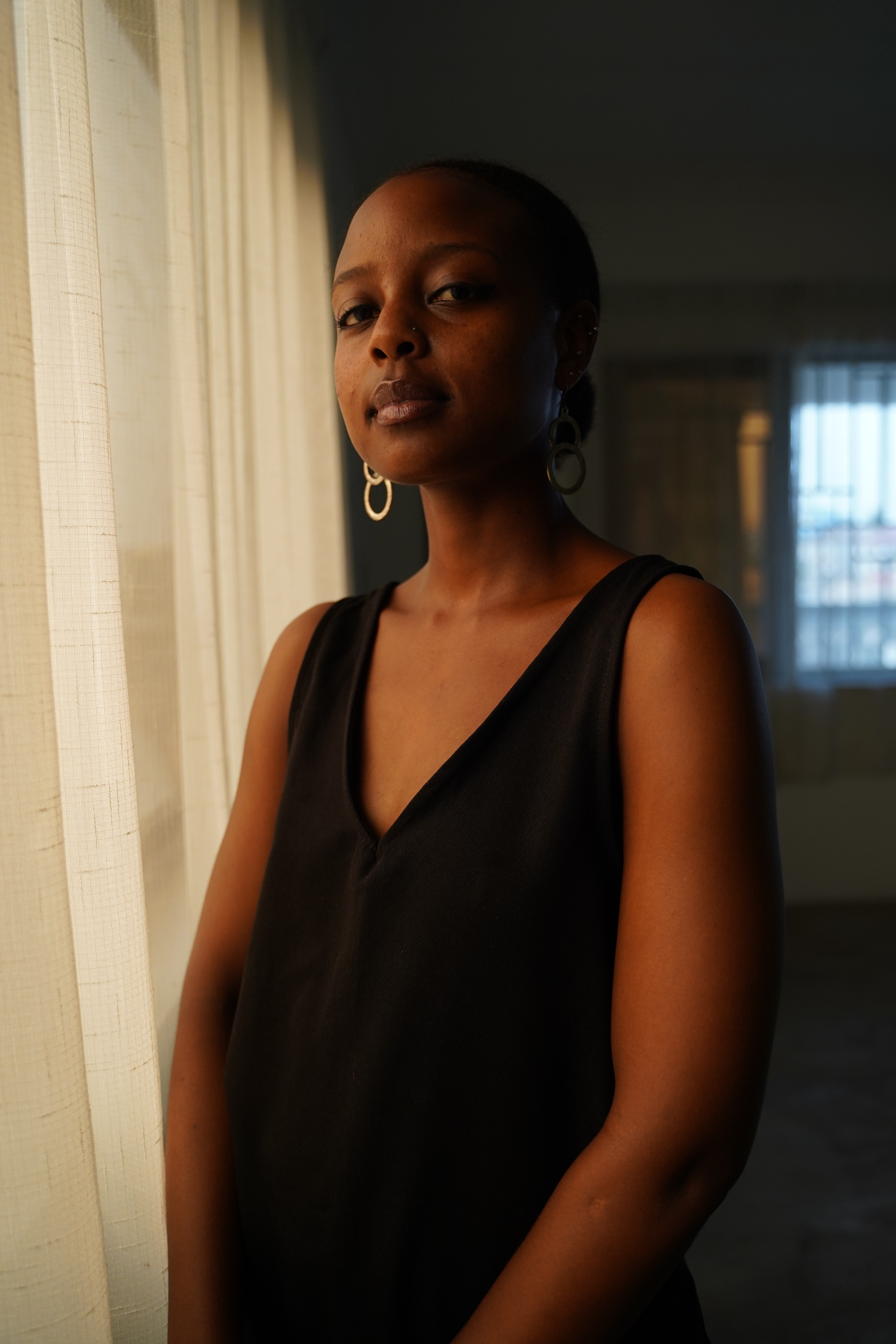 Dark-skinned woman in a black top and hoop earrings stands by a sheer curtain, looking at the camera.