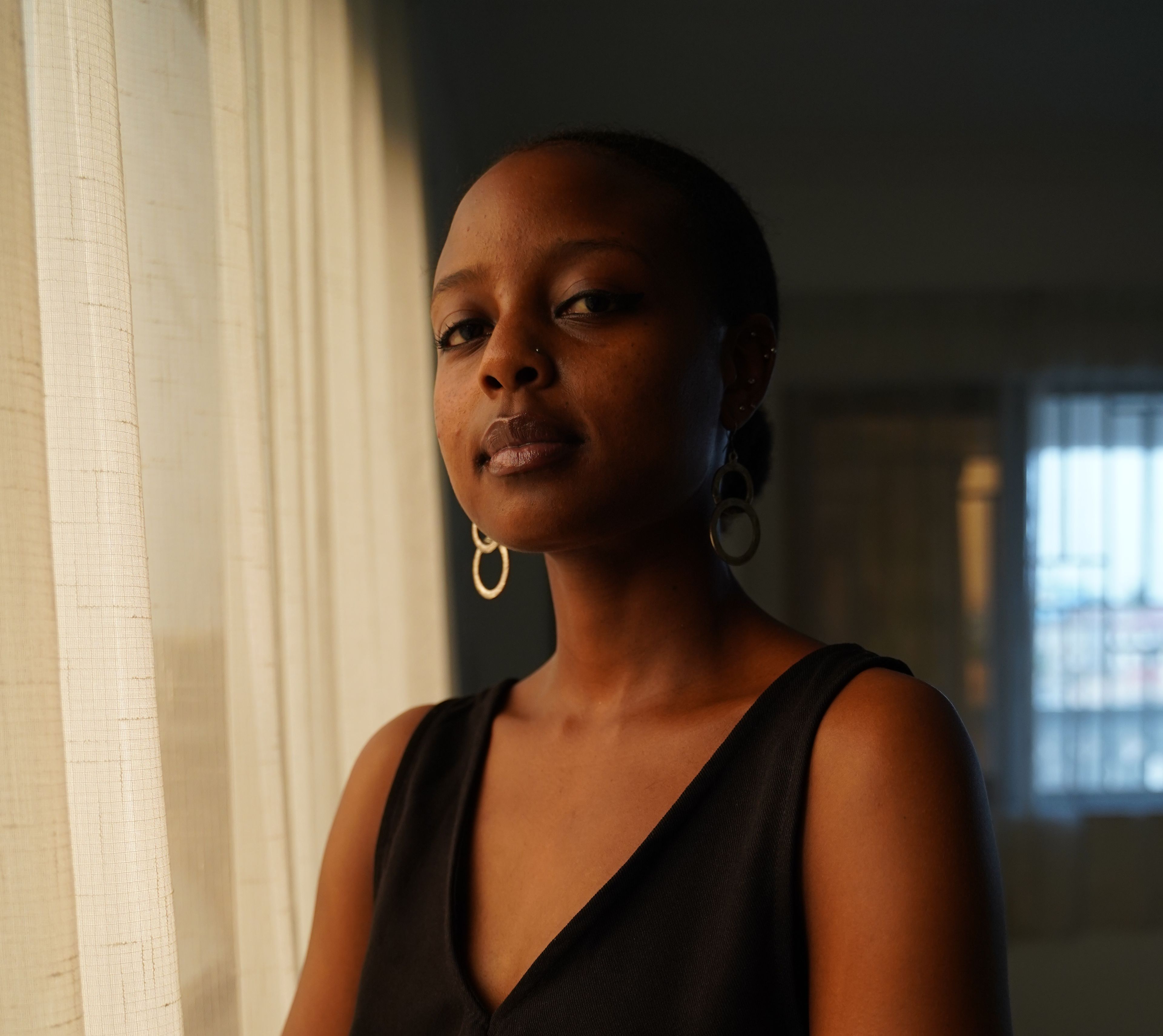 Dark-skinned woman in a black top and hoop earrings stands by a sheer curtain, looking at the camera.