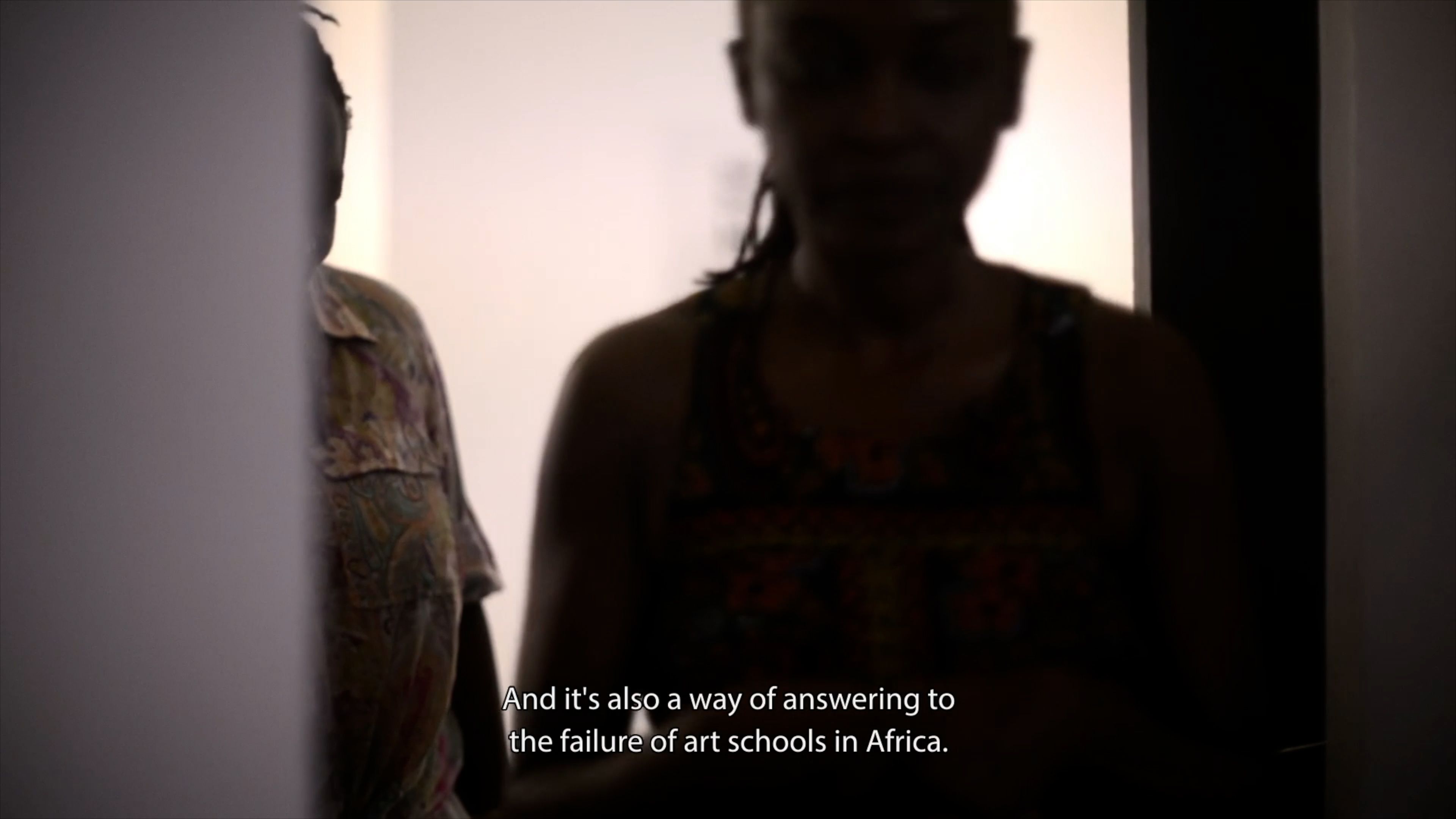 Two people are partially visible in a dimly lit room, their faces obscured by shadow. White text across the bottom reads: "And it's also a way of answering to the failure of art schools in Africa."