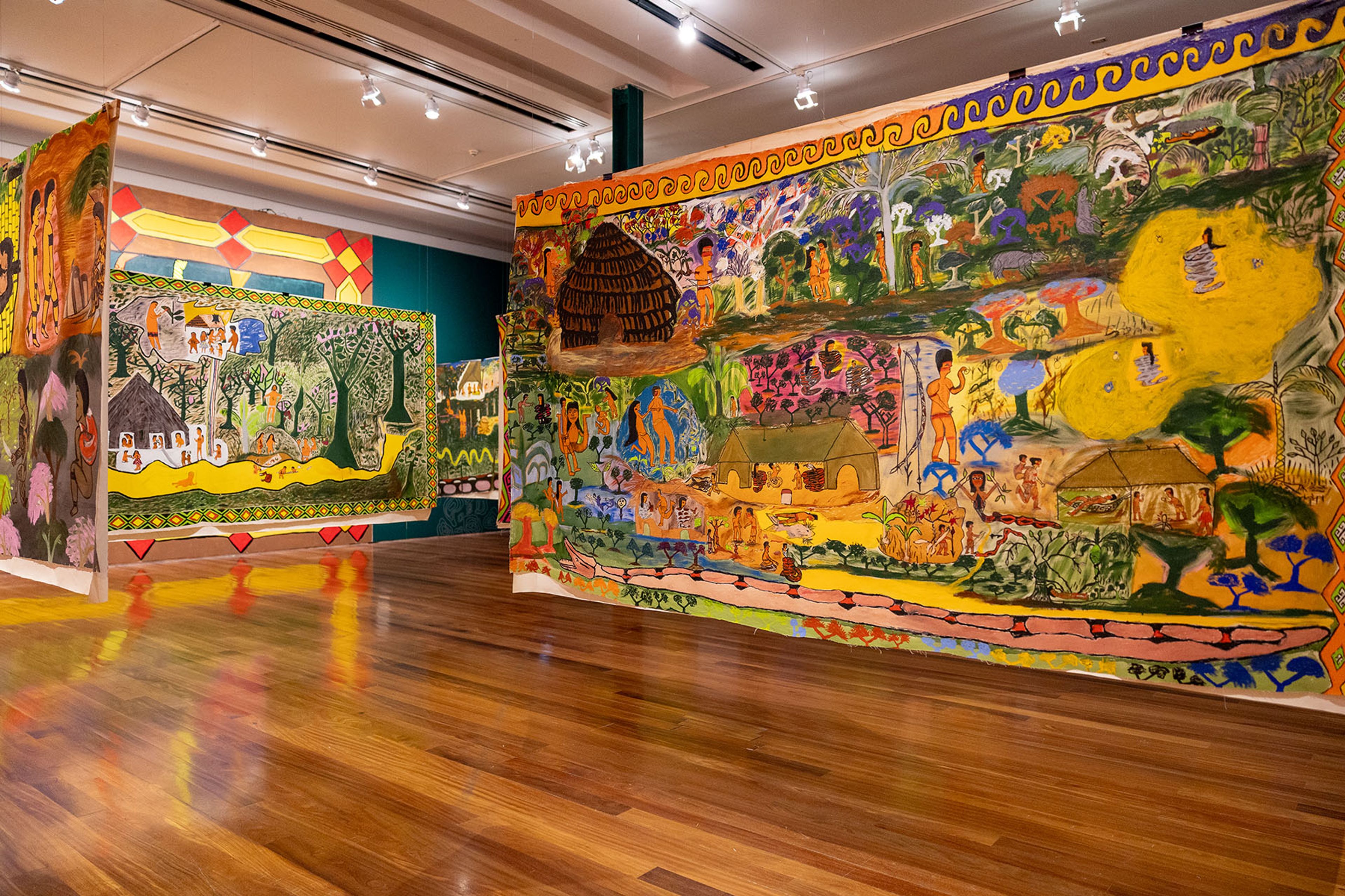 Several large, colorful folk-art murals depicting village scenes and nature are displayed in a brightly lit gallery with a polished wooden floor.