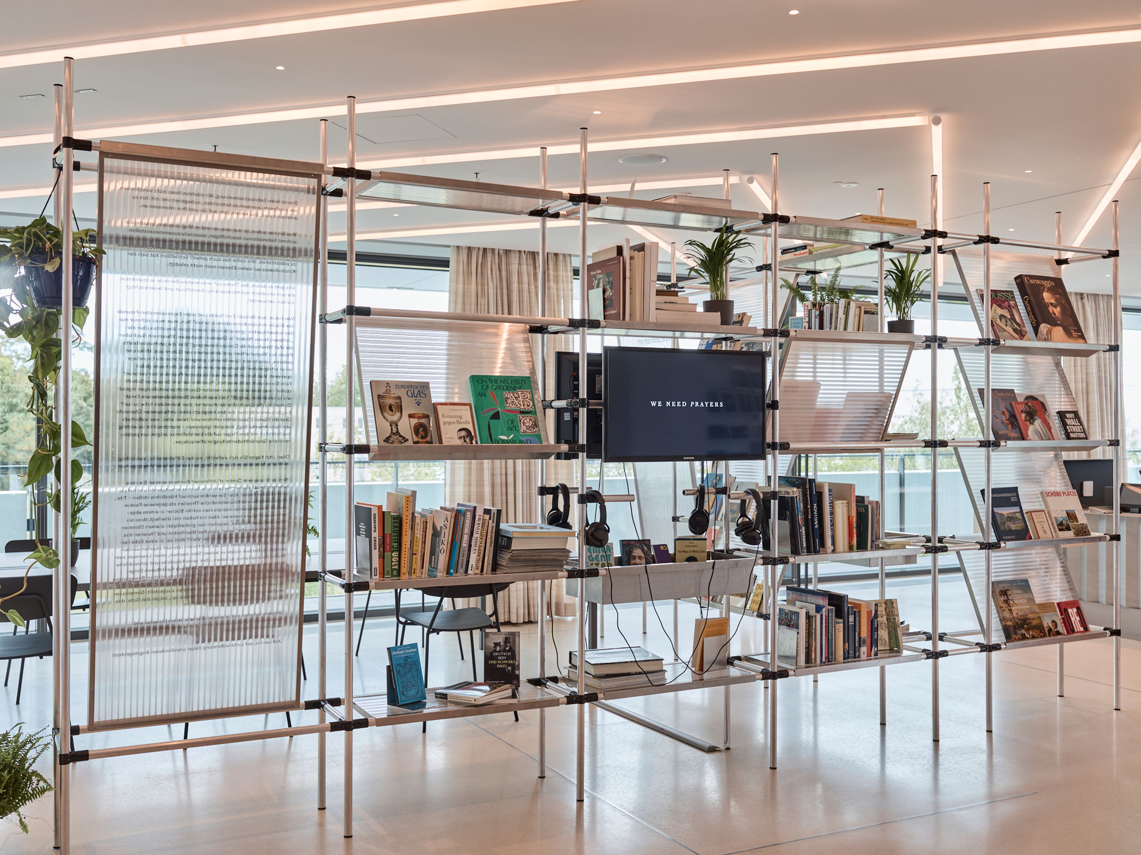 A modern modular shelving unit with books, plants, a TV, headphones, and a translucent panel, set in a bright, contemporary interior.