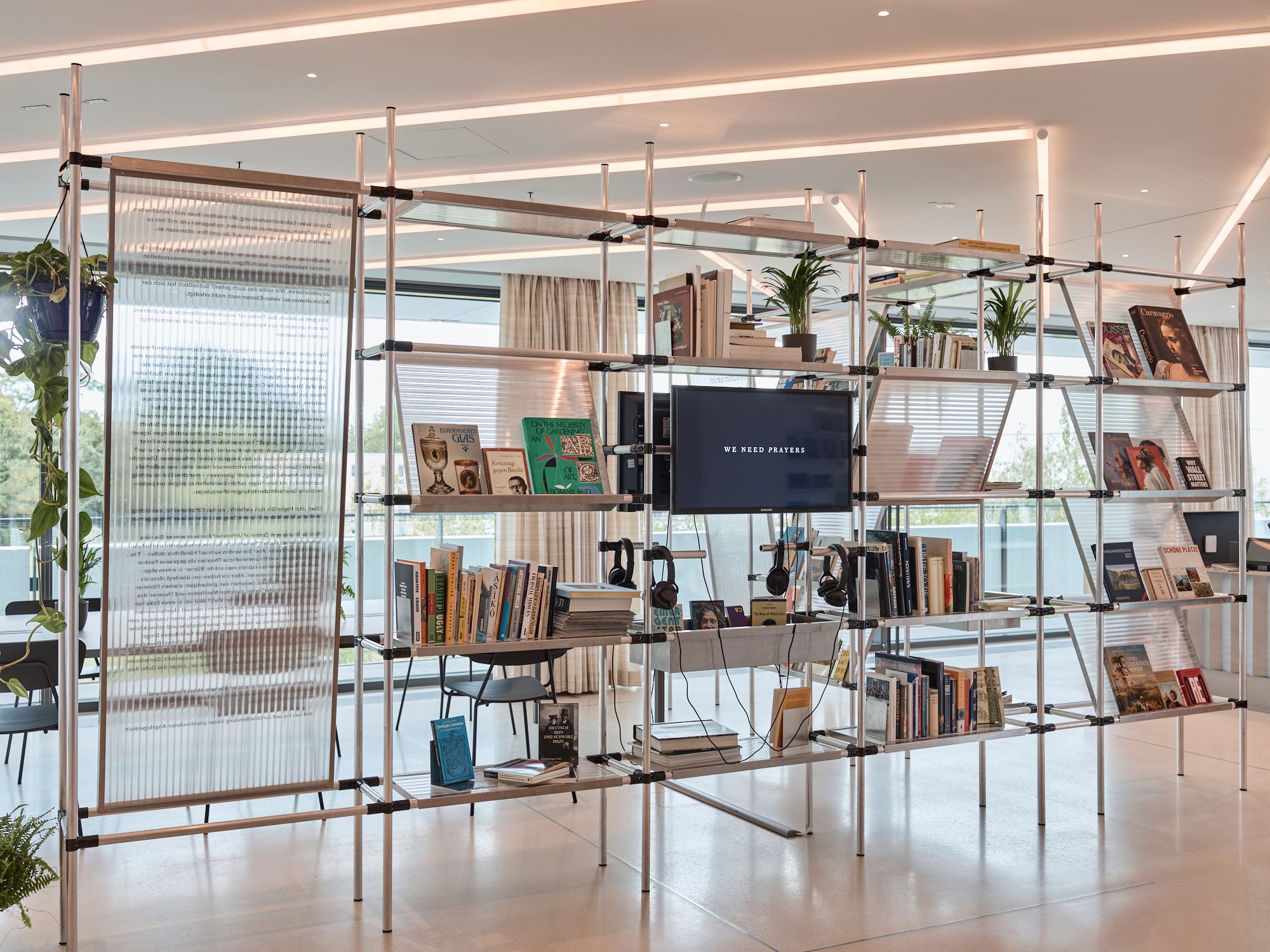 A modern modular shelving unit with books, plants, a TV, headphones, and a translucent panel, set in a bright, contemporary interior.