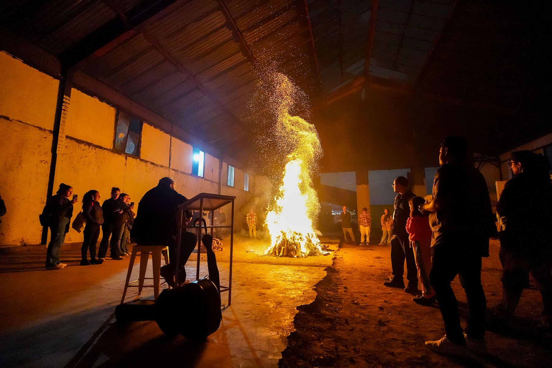 People gather around a large bonfire with flying sparks in a dark indoor space.