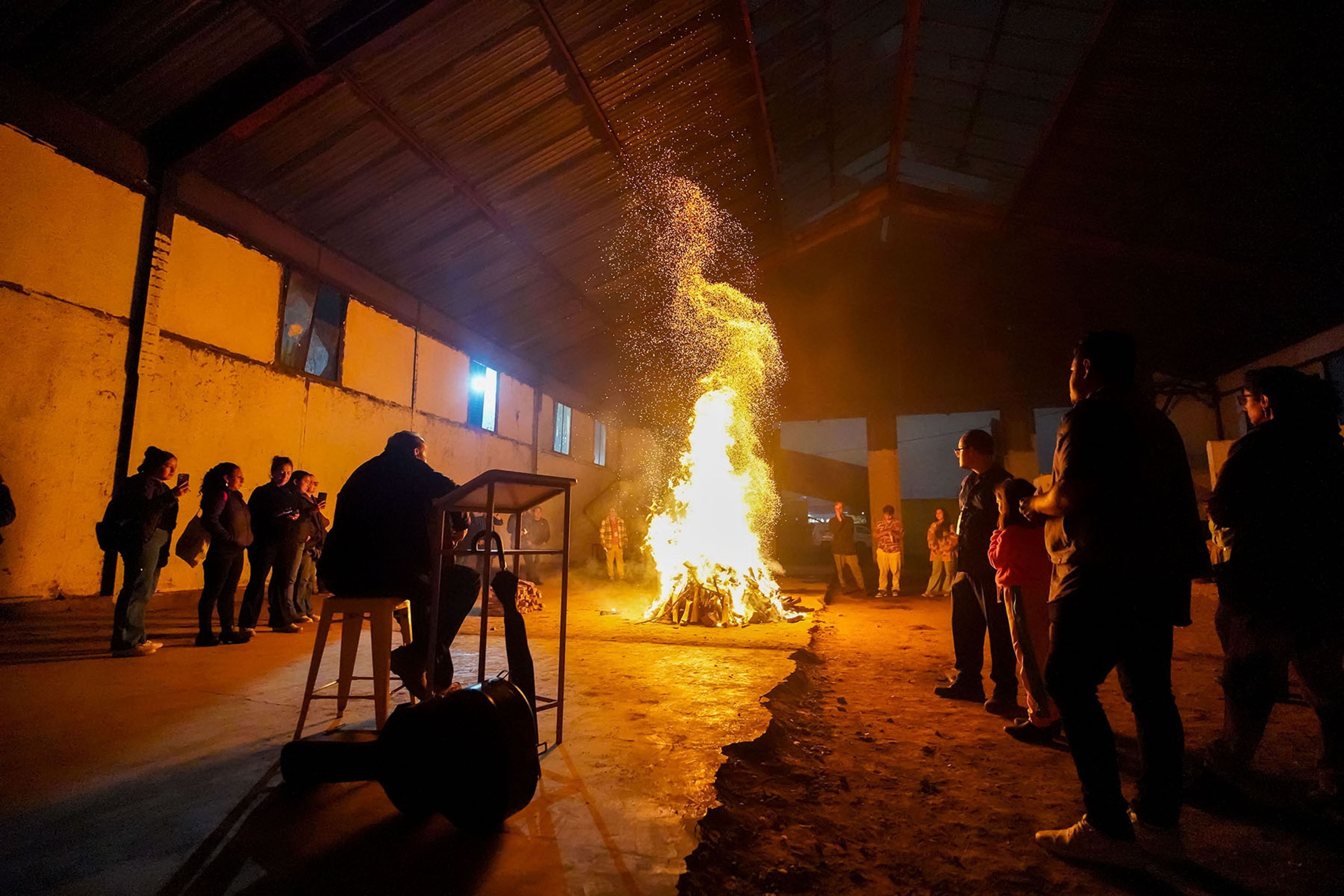 People gather around a large bonfire with flying sparks in a dark indoor space.