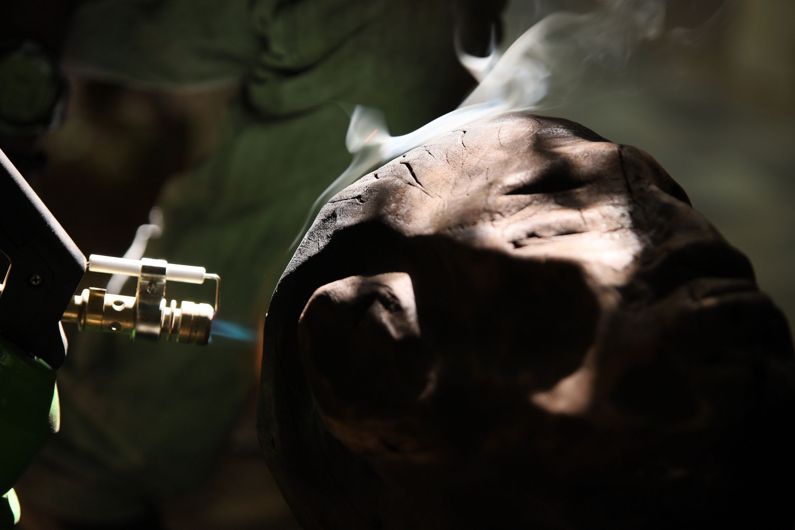 A close-up of a blue blowtorch flame heating a dark, textured piece of wood or material, causing smoke to rise.