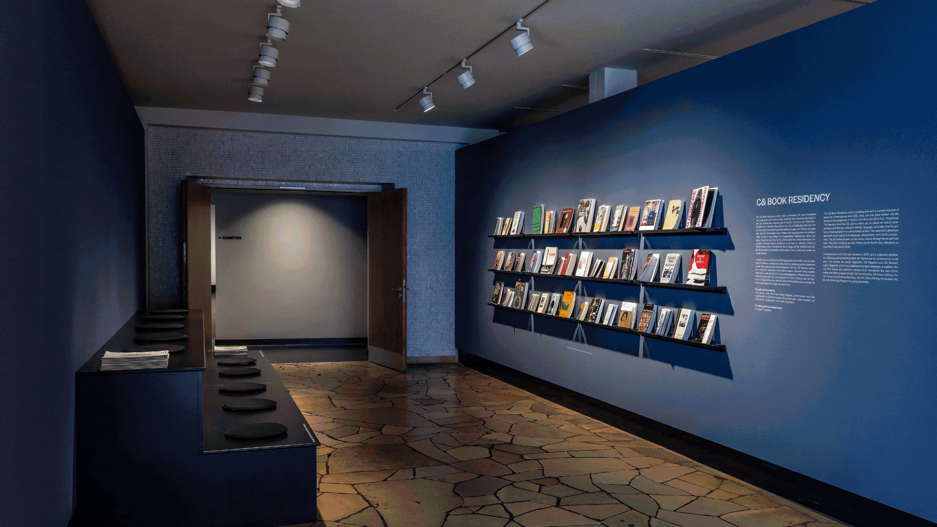 An exhibition space with a blue wall featuring books on shelves and text about a 'GB BOOK RESIDENCY', a dark counter on the left, and a doorway.