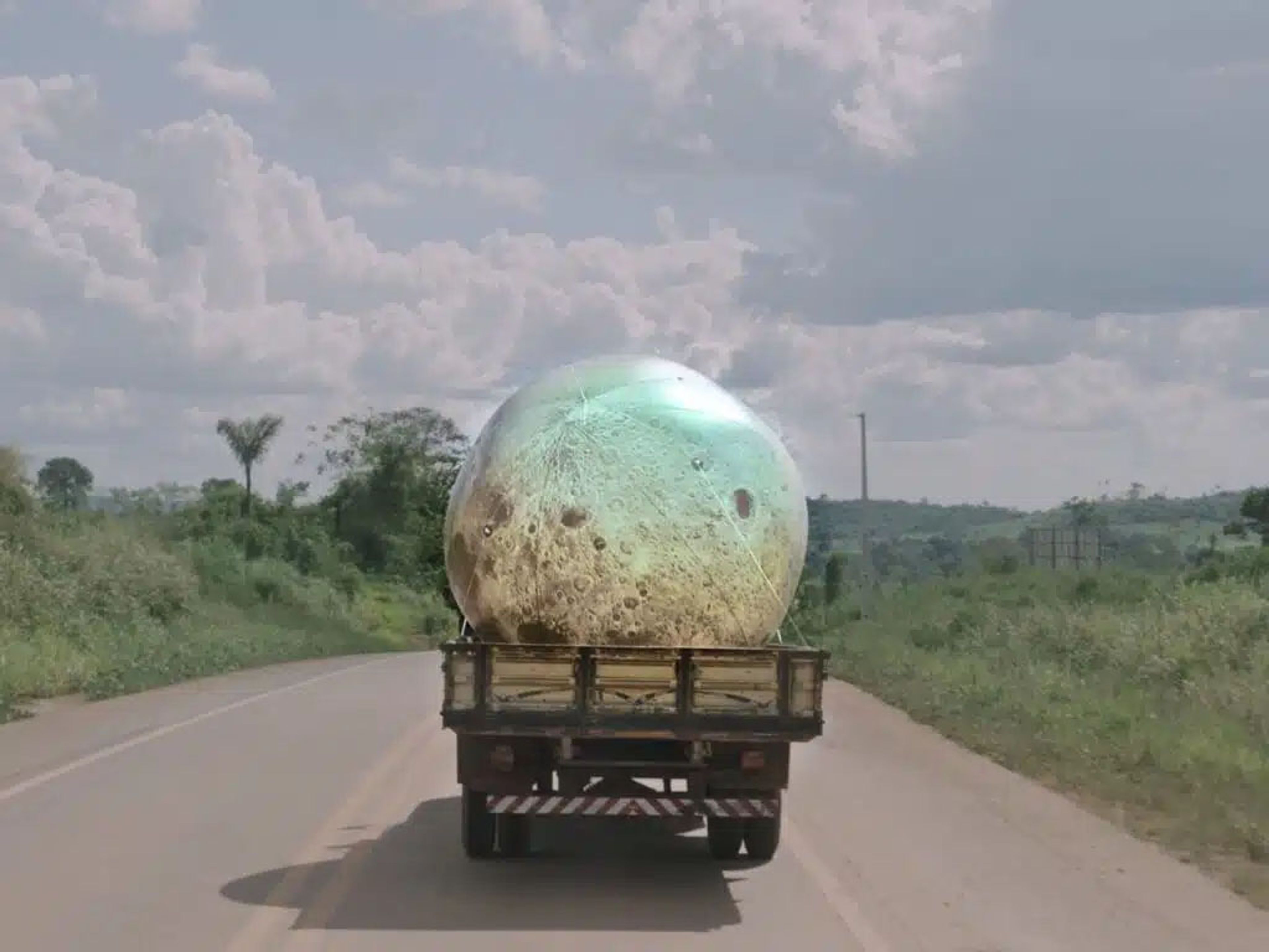 A truck drives down a road carrying a giant, glowing, cratered sphere resembling the moon.