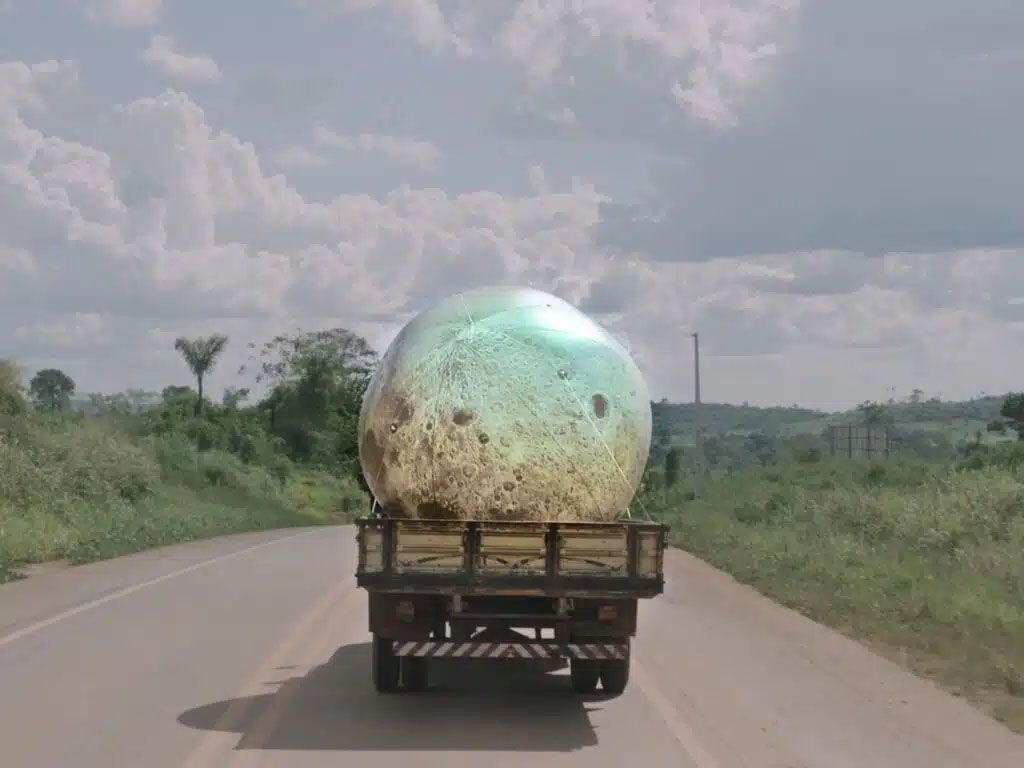 A truck drives down a road carrying a giant, glowing, cratered sphere resembling the moon.