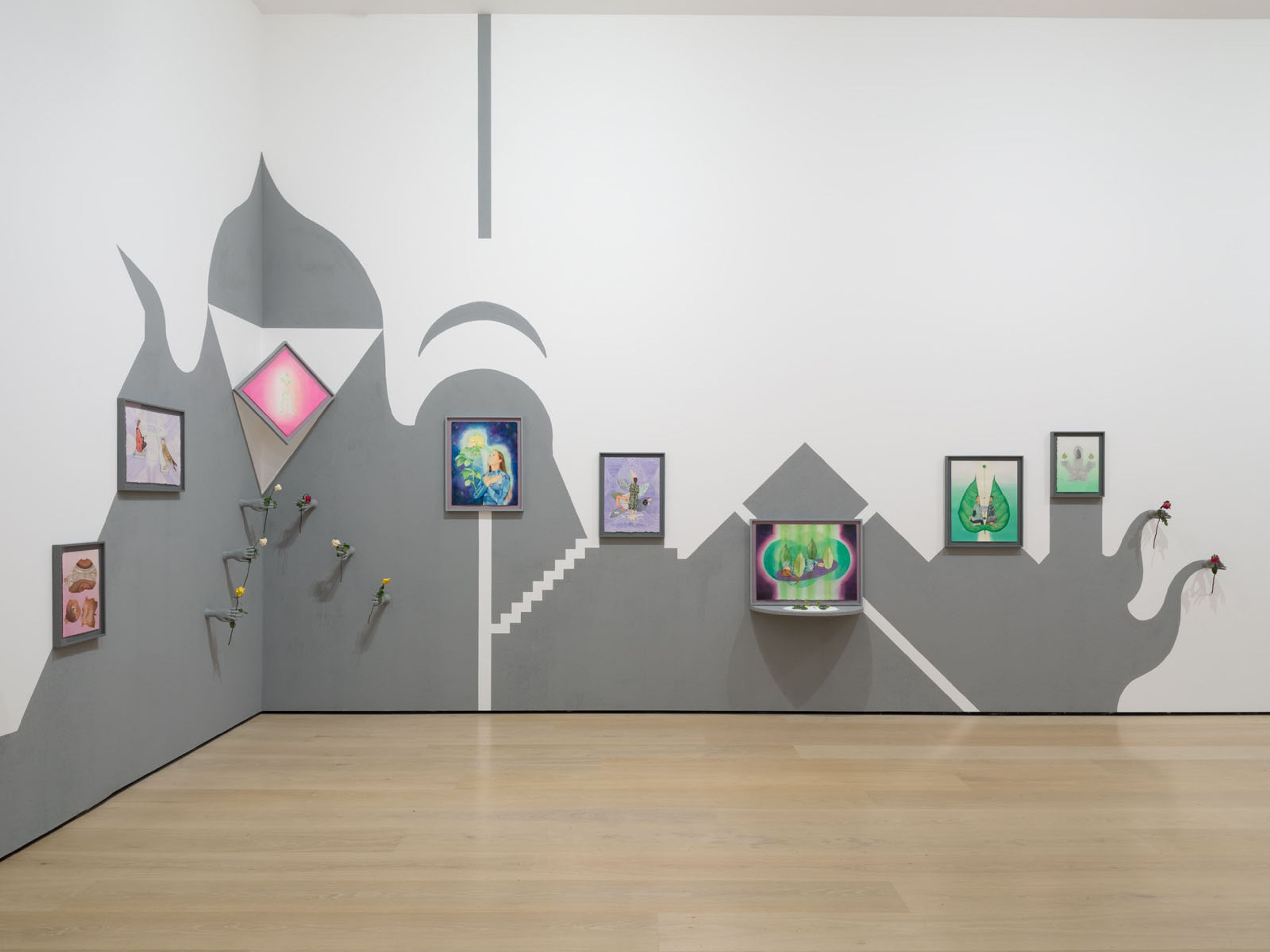 A gallery installation across two walls featuring gray and white abstract painted designs, multiple framed artworks, a TV screen, and attached elements like flowers.