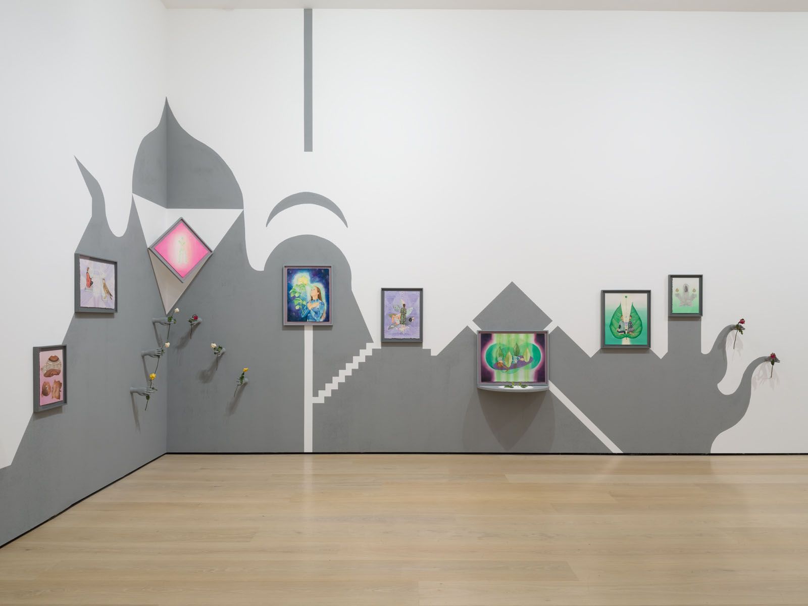 A gallery installation across two walls featuring gray and white abstract painted designs, multiple framed artworks, a TV screen, and attached elements like flowers.