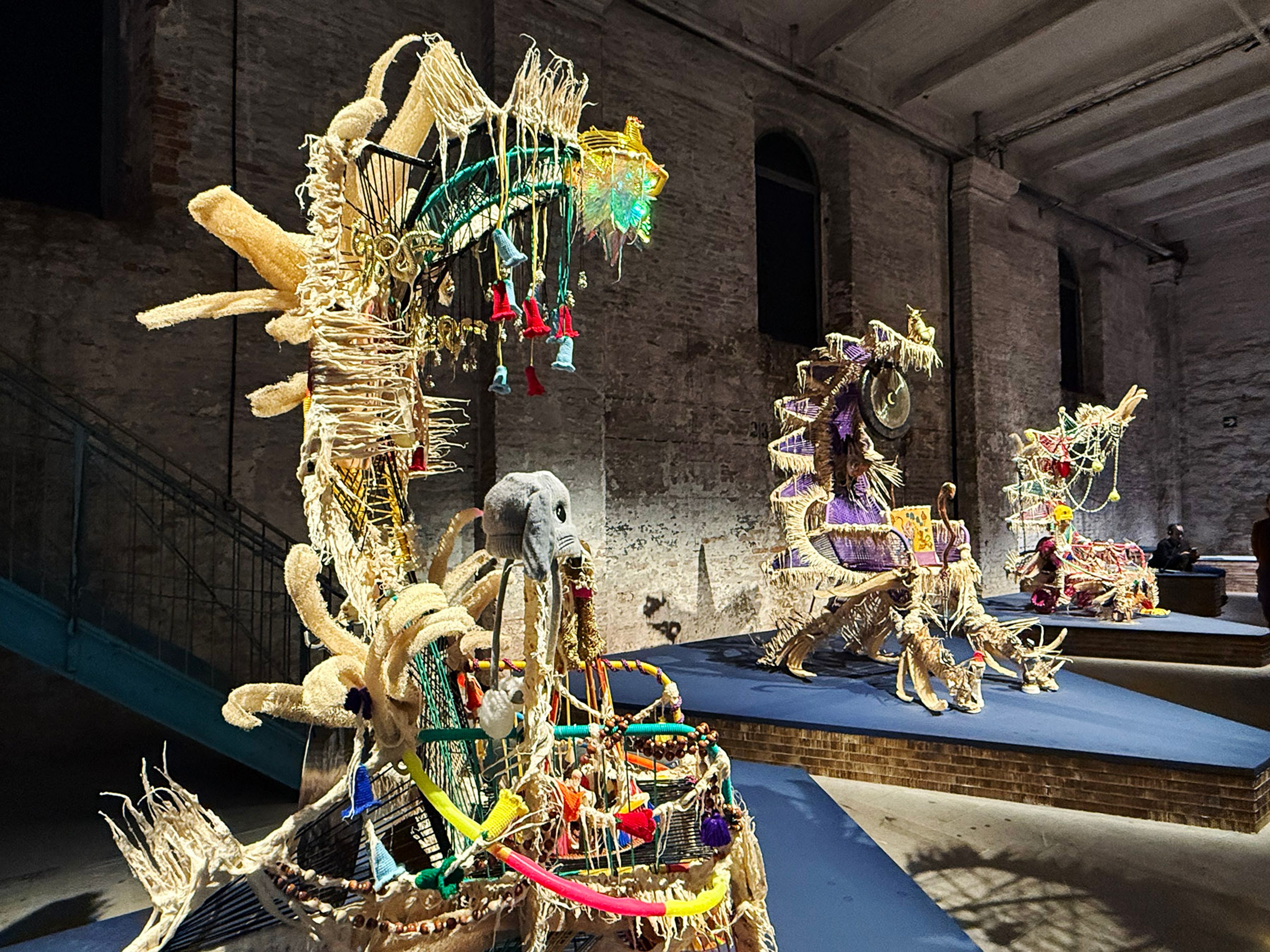 Three elaborate, colorful textile and mixed-media sculptures resembling mythical creatures, displayed on blue platforms in a brick-walled gallery.