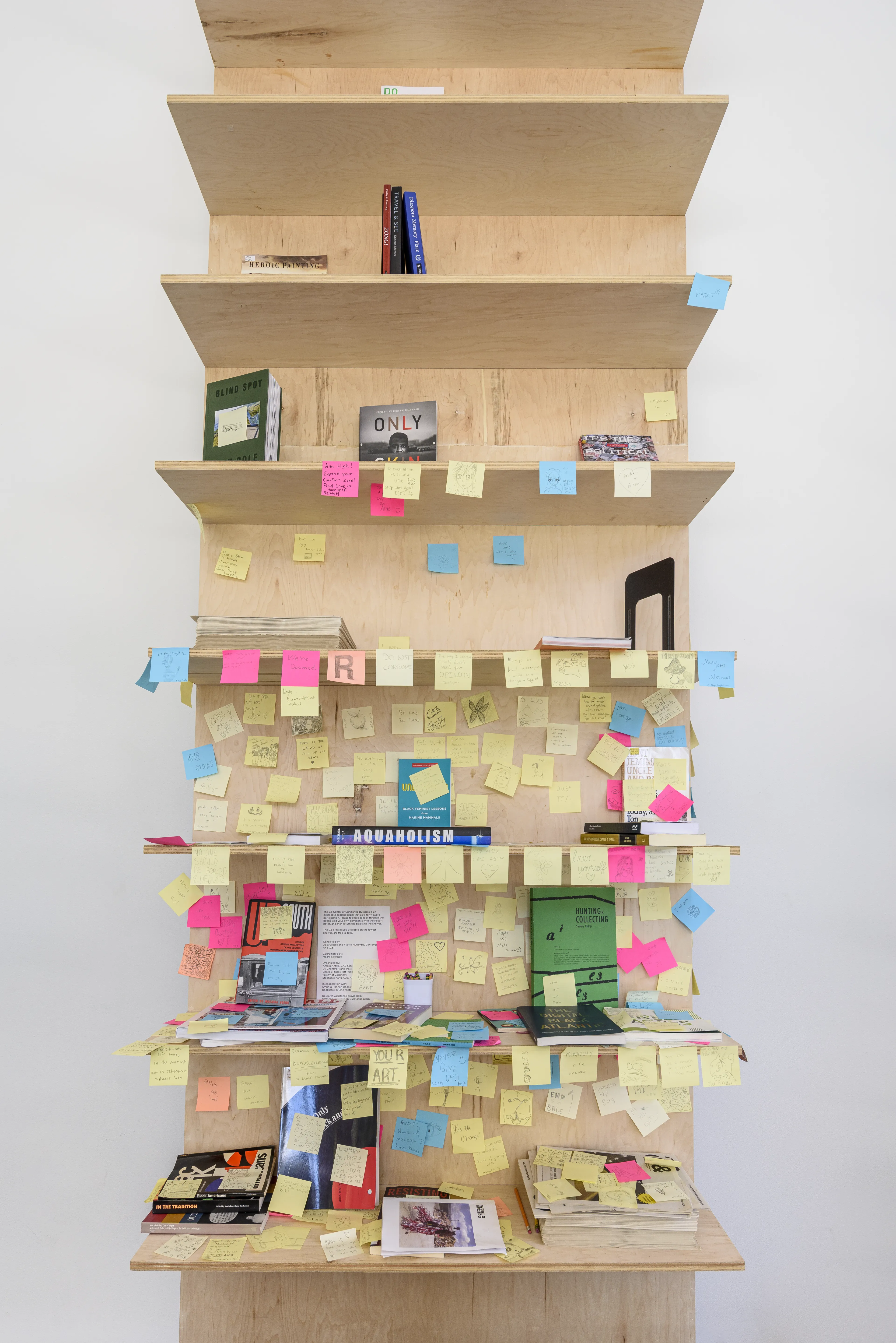 A tall, light wood bookshelf densely covered with books, magazines, and yellow, pink, and blue sticky notes.